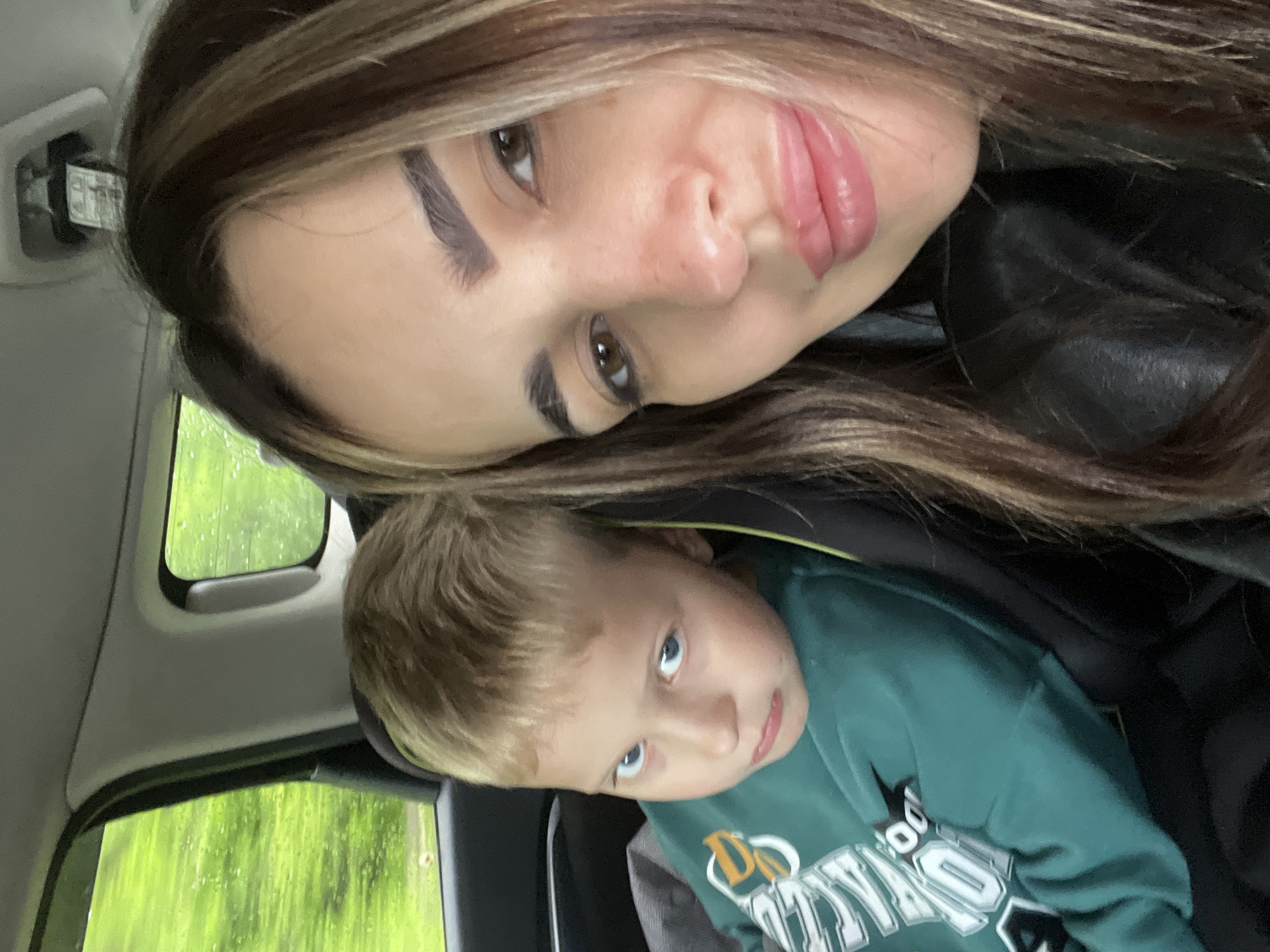 An adult female and a young male child are seated inside a vehicle. The female, positioned on the right, has long, multi-toned brown hair, brown eyes, defined eyebrows, and full, glossy pink lips, looking directly at the camera. She wears a dark garment, possibly a jacket. The child, situated in a dark car seat on the left, has short blonde hair and blue eyes, and is wearing a long-sleeved green sweatshirt with white and yellow design elements. He looks towards the camera with a slight frown. Visible through the vehicle's windows are raindrops and an expansive, bright green natural landscape, indicating an outdoor, possibly vegetated, environment. The interior of the vehicle, including portions of the headliner and window frames, is visible. The scene takes place in the city of Unknown, Unknown.