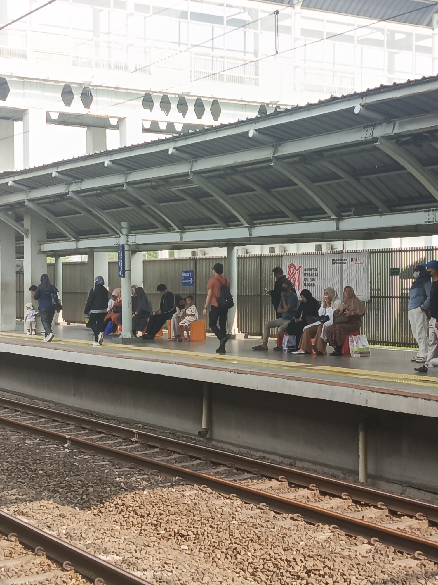 A street-level, eye-level, medium shot depicts a train station platform with people waiting. The scene is located at the Telagamurni train station in Telagamurni, Indonesia. A covered platform with a corrugated metal roof, supported by white pillars, extends horizontally across the image. Beneath the roof, people are seated and standing, awaiting transportation. The platform features a yellow line demarcating the edge. In the background, a multi-story building with a glass facade rises above the station. Several individuals are visible on the platform: a person in an orange shirt walks right, facing away from the camera; a group of people, including women wearing hijabs and men, are seated on a bench and chairs; a child in a white shirt and shorts stands near the edge of the platform. A young child in a white shirt and light-colored shorts stands near a woman in a blue jacket and dark pants, who is also facing away from the camera. Another woman in a black hijab and dark clothing stands on the platform to the left, facing the camera. Railway tracks, filled with gravel, are visible in the foreground, with two sets of parallel tracks running horizontally. A banner with text and an image is affixed to a fence in the background. The lighting suggests it is daytime, with bright, diffused light illuminating the scene.