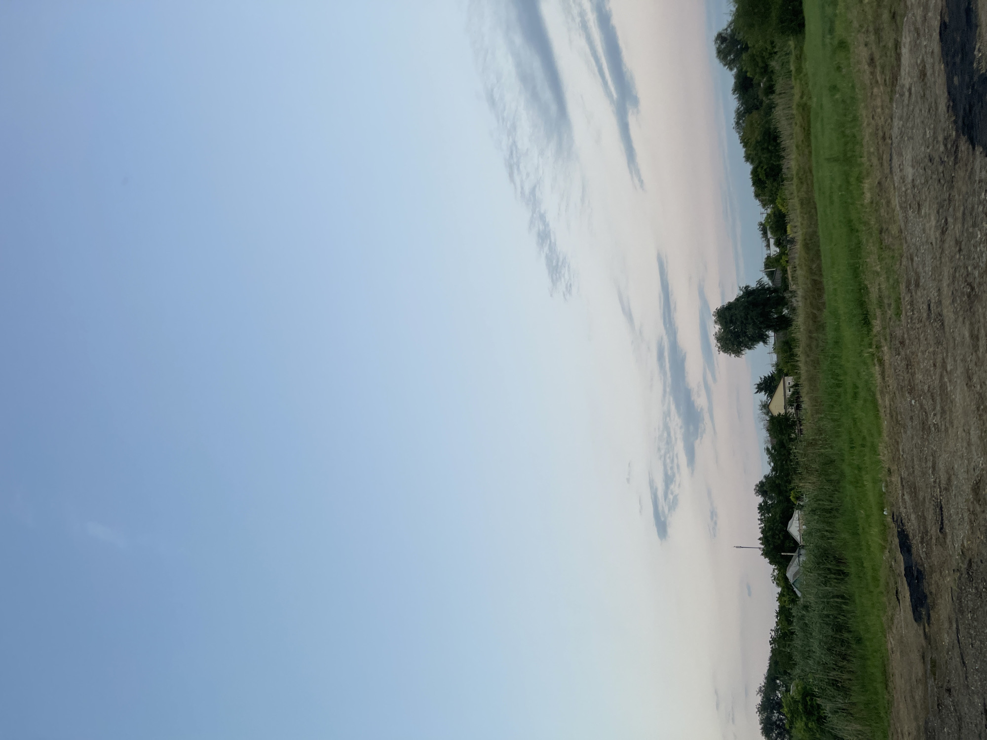 This is a photograph of a rural landscape with a clear sky. The sky is a gradient of light blue to pale pink, with thin, wispy clouds towards the horizon. Below the sky is a band of green grass and vegetation, with several small houses and trees visible in the middle ground. In the foreground, there is a dirt path with some dark, possibly tar-like, patches. The scene appears to be a quiet, undeveloped area.