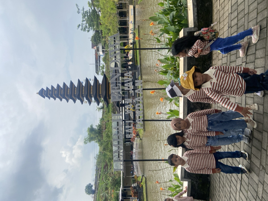 Five children stand side-by-side, wearing striped shirts and jeans, posing in front of a pond. Behind them is a towering black temple-like structure, a sign that reads "UMBUL BALI VAN JAVA PANDAAN", and lush greenery. The sky is overcast. The children are on a paved pathway, with orange flowers scattered throughout the landscape.