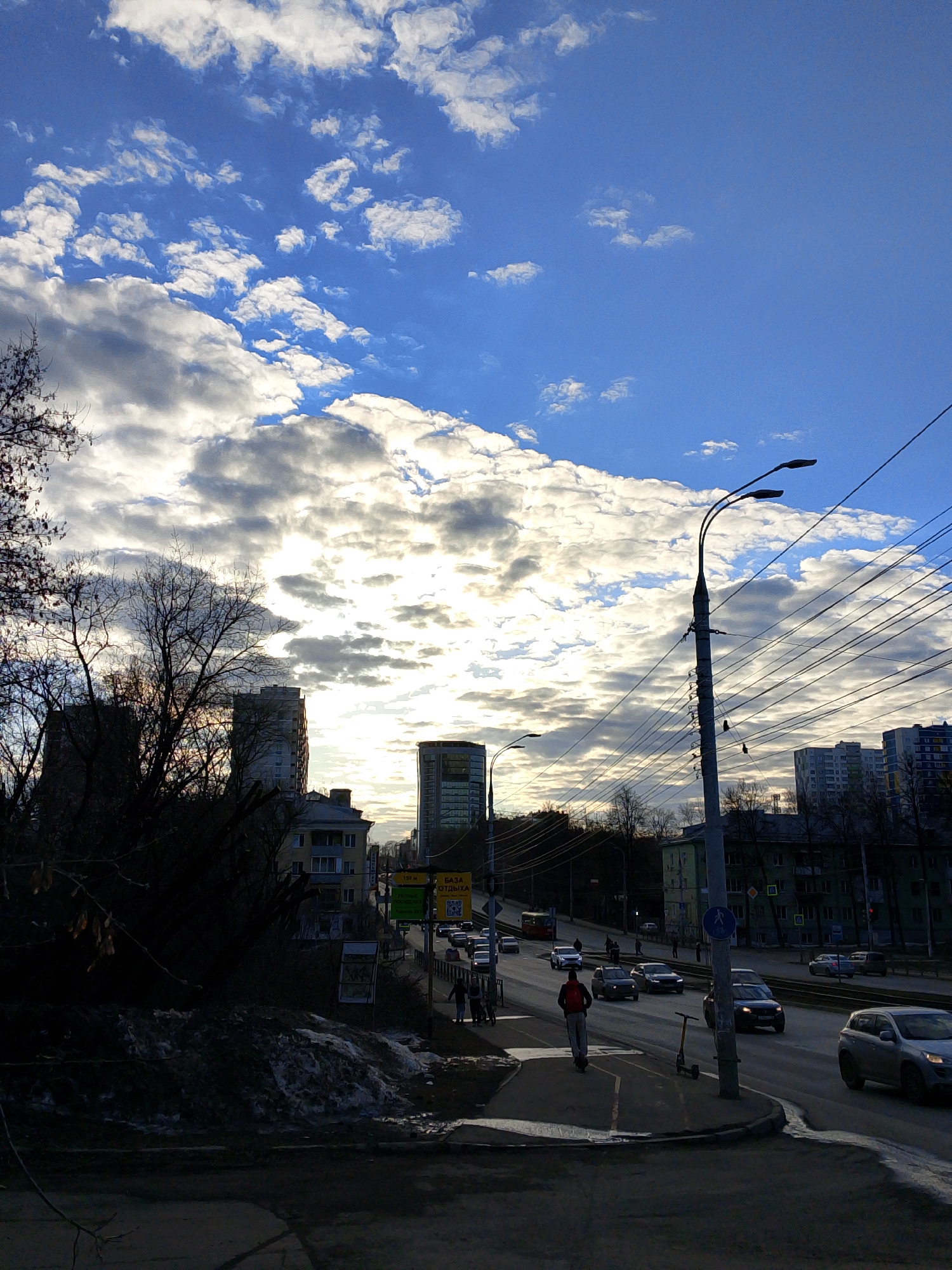 The image depicts a street scene in Izhevsk, Russia. The sky is a mix of blue and white clouds, with the sun obscured behind the clouds, creating a bright glow. Below the sky, there are buildings of varying heights and architectural styles, interspersed with bare trees. A street with traffic occupies the middle ground, with several cars and at least one person on an electric scooter visible. A pedestrian is also seen walking on the sidewalk. Streetlights and utility poles with numerous wires run along the right side of the street. On the left, there is a snow bank with some visible vegetation. The foreground is dark, suggesting that the photo was taken from a slightly elevated position. A sign with Cyrillic characters is visible on the side of the road.