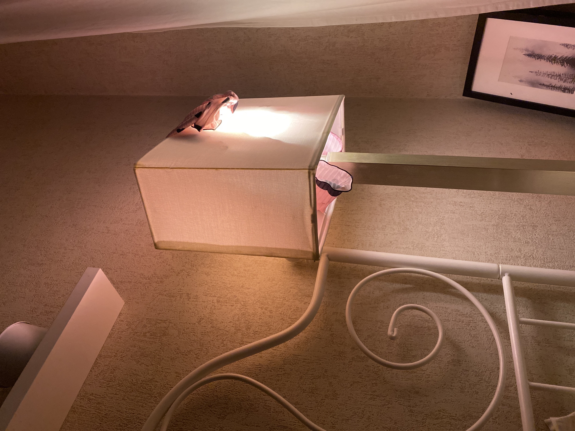 The image depicts a bedroom interior, possibly in a residential building. The main subject is a bedside lamp with a rectangular shade, emitting a warm glow. The lamp is attached to a metal arm extending from the wall. The room has textured wallpaper in a beige or light brown tone. In the upper right corner, there is a framed picture with a dark frame and a white mat. A white wrought-iron bed frame is partially visible, with its decorative curves. Also visible are lingerie items draped over the lamp shade.