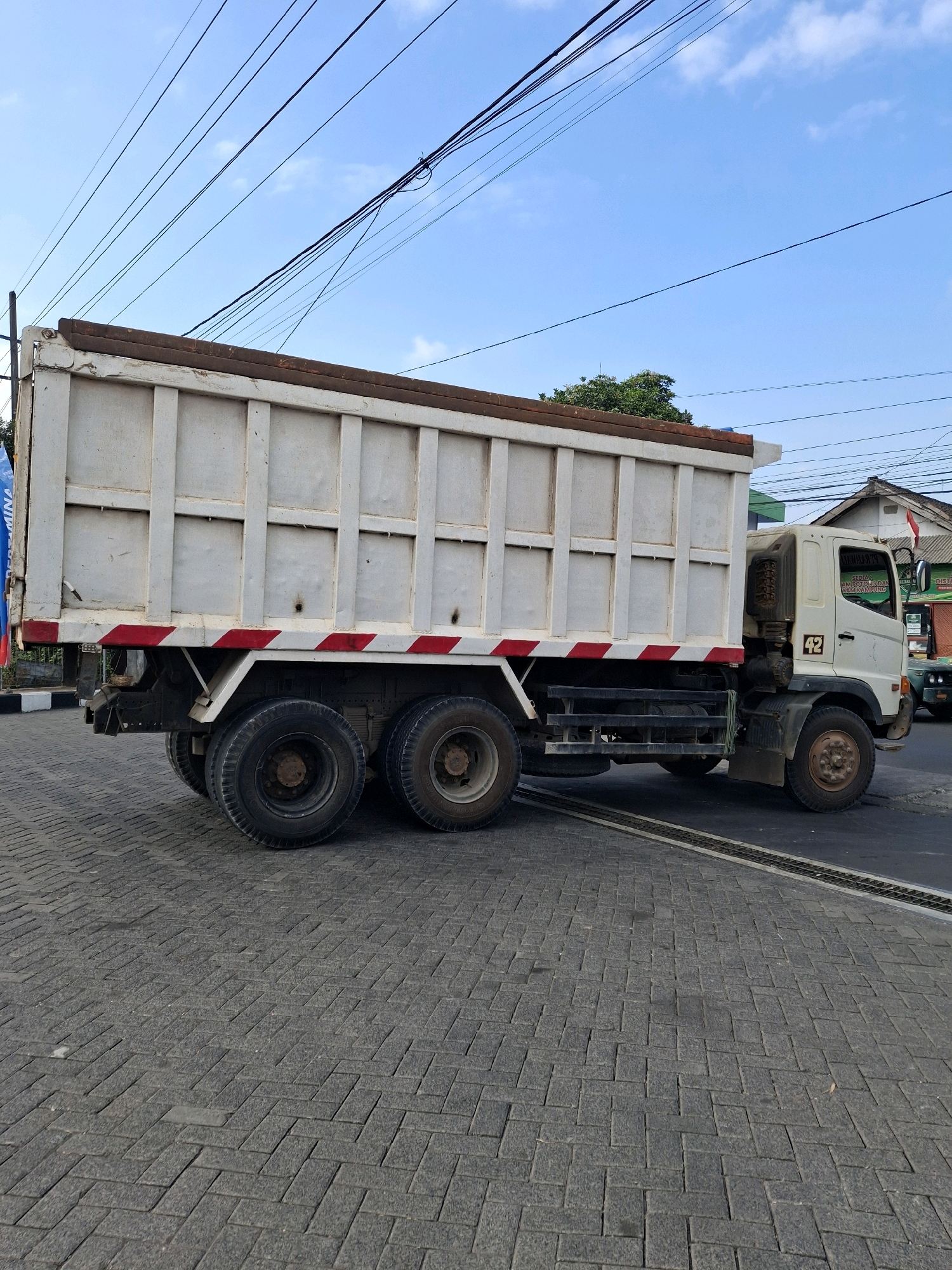 A side view of a white dump truck is captured on a paved street in Lumajang, Indonesia. The truck has a large, weathered white dump bed with visible reinforcing beams. Red and white reflective stripes adorn the lower side of the dump bed. The truck has dual rear wheels on each side and single front wheels. The front cab is also white and shows some wear and tear, with the number "42" visible on the driver's side. A green and white sign with text is visible on the side of the truck cab. In the background, overhead power lines crisscross the bright blue sky with scattered white clouds. A building with a red and white flag is visible to the right of the truck, along with trees and other vehicles. The ground is paved with interlocking grey bricks.