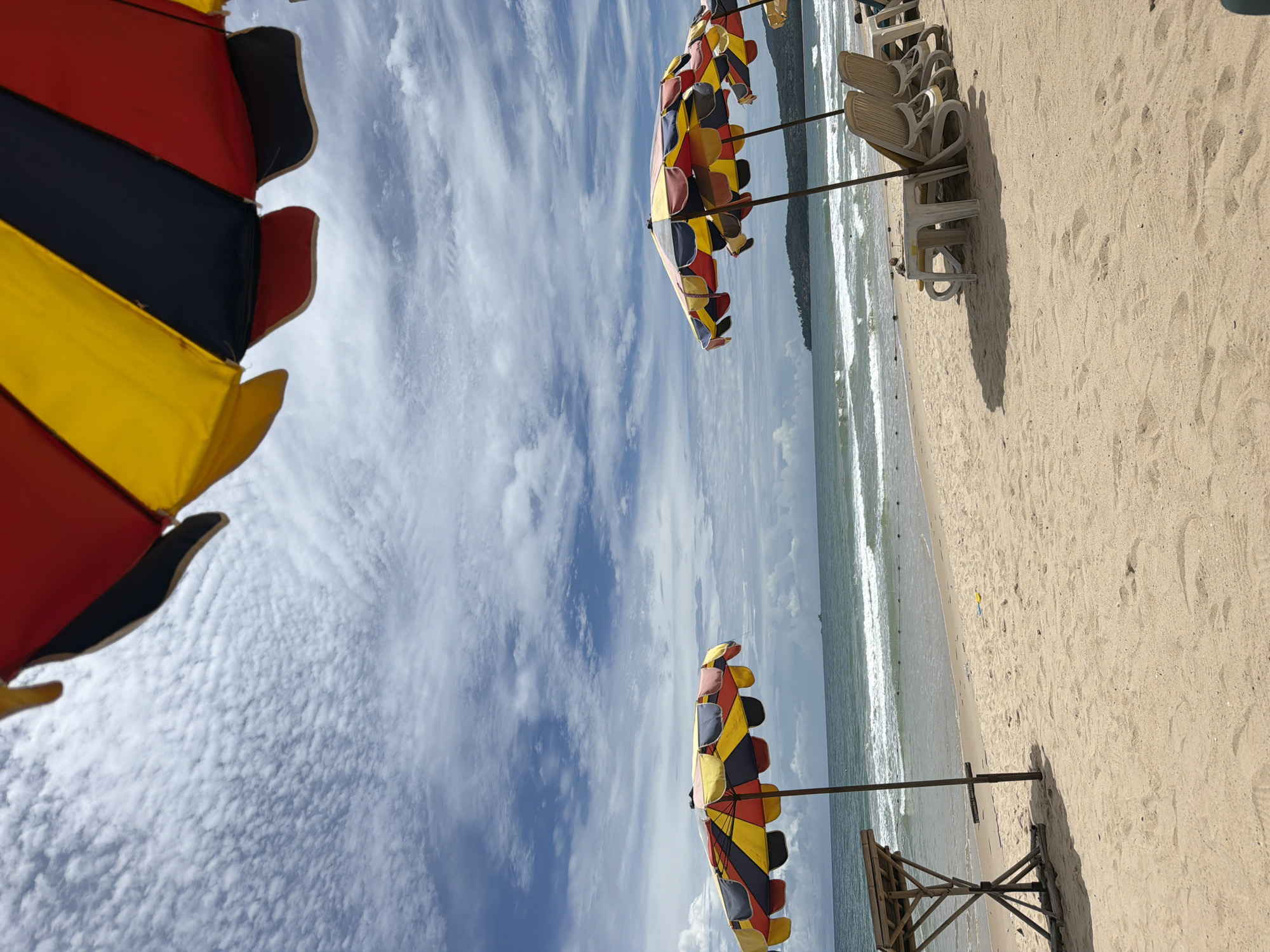 This is a full, eye-level shot of a beach in Choeng Thale, Thailand. In the foreground, there is a sandy beach with several beach chairs and umbrellas. The umbrellas are colorful, with alternating red, yellow, and blue panels. In the middle ground, there is the ocean with gentle waves and a faint horizon line. In the background, there are rolling hills covered in green foliage. The sky is partly cloudy with patches of blue sky. The overall impression is one of a serene and beautiful beach scene on a pleasant day.