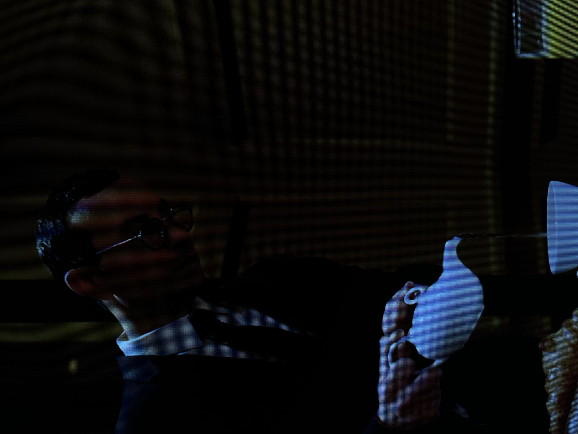 A man wearing a dark suit and glasses is pouring liquid from a white teapot into a white cup. The man is partially obscured by darkness, but his head and upper body are visible, as is his left hand holding the teapot. The background is dark, with some hints of wooden paneling. There is also a glass of yellow liquid visible to the right of the cup. The scene is likely indoors, possibly in a restaurant or dining room. There is no information in the image to suggest the location is in Łódź, Poland.