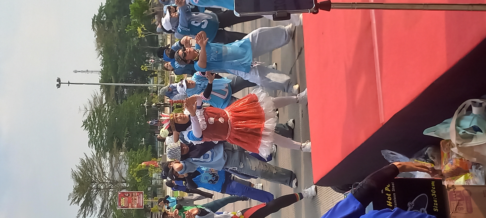 In Melayu, Indonesia, a group of people are engaged in an outdoor exercise routine. The scene is set in a paved public space with trees and streetlights visible in the background. In the foreground, a bright red stage or platform is present.
The central figure, a woman wearing a distinctive red and white outfit with a feathered headdress, leads the group in a choreographed dance. She is wearing a red dress with ruffled white trim at the neck and hem, and white leggings. Her hands are held in front of her chest in a preparatory pose.
Surrounding her, several other individuals, mostly women, are dressed in similar activewear, predominantly in shades of blue and grey. Many are wearing hijabs. They are all performing synchronized movements with their arms and legs, indicating a coordinated exercise class or performance. Some are wearing athletic shoes, while others have on casual footwear.
To the left of the frame, a dark blue bag with "Hot P" and "Stainless Steel" printed on it is visible, along with some packaged goods. A hand is reaching into the bag. The overall atmosphere suggests a community event or a public fitness gathering. The lighting indicates it is daytime, with sunlight casting shadows.