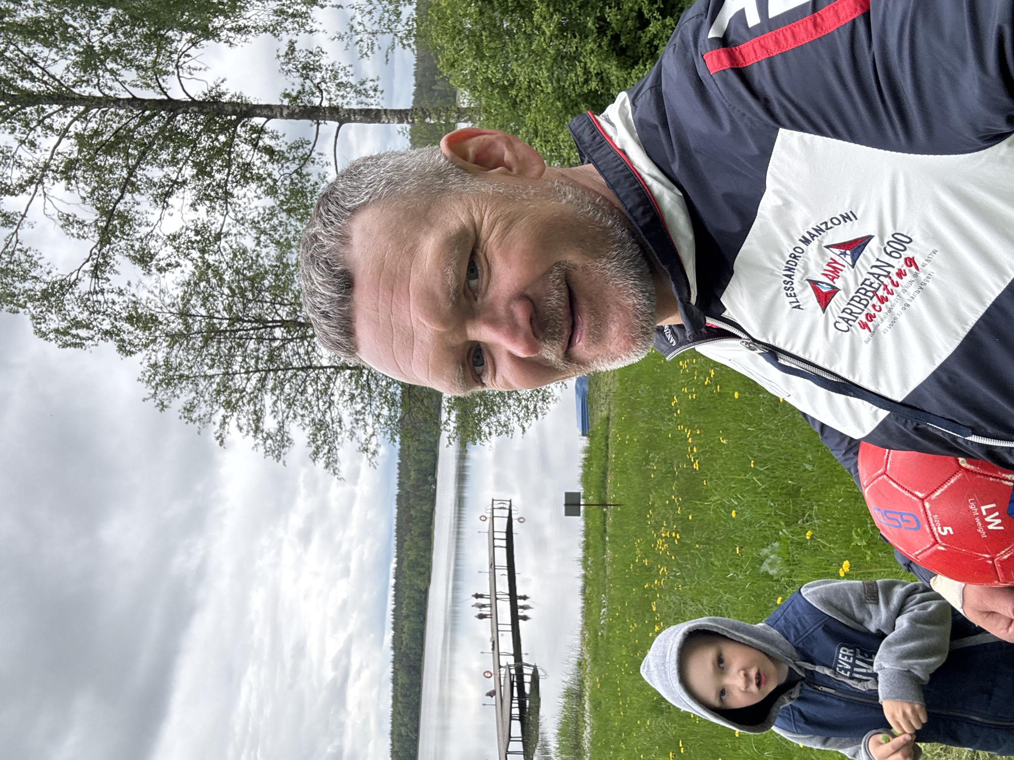 A man and a young child stand in a grassy field near a lake with a wooden pier. The man is wearing a jacket with the words "ALESSANDRO MANZONI," "CARIBBEAN 600 yachting," and the coordinates "Bout Warrior Latst Positio 017 30.665N 06 46.817W" on it. The child is wearing a hooded jacket. A red soccer ball is visible, marked with "GS" and "LW" for "Light weight." The location, based on the jacket's coordinates, is near Aleksandrovka, Russia. The sky is overcast, and trees line the opposite shore of the lake.