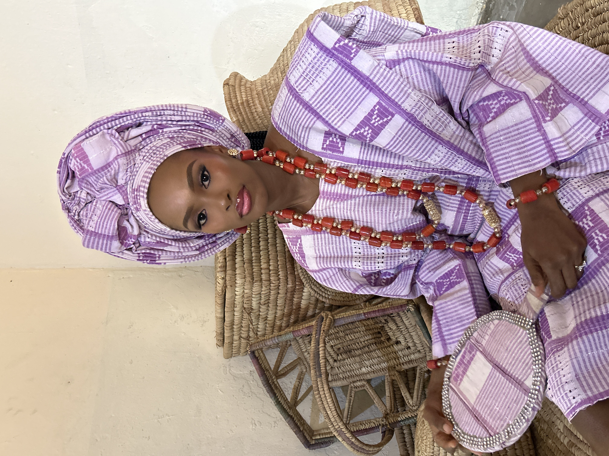 A woman wearing a purple and white striped outfit and headscarf sits in a basket chair, holding a decorative mirror. The outfit has a traditional African design with intricate patterns. She wears red beaded necklaces and bracelets, and her makeup is flawless, with bold eyeliner and pink lipstick. The background consists of woven baskets, suggesting an indoor setting or a marketplace. The lighting is soft, implying it could be daytime. The woman's expression is calm and poised. The mirror she holds is oval-shaped and also features a purple and white checkered pattern, with a border of what appears to be rhinestones. The overall impression is one of elegance and cultural pride.