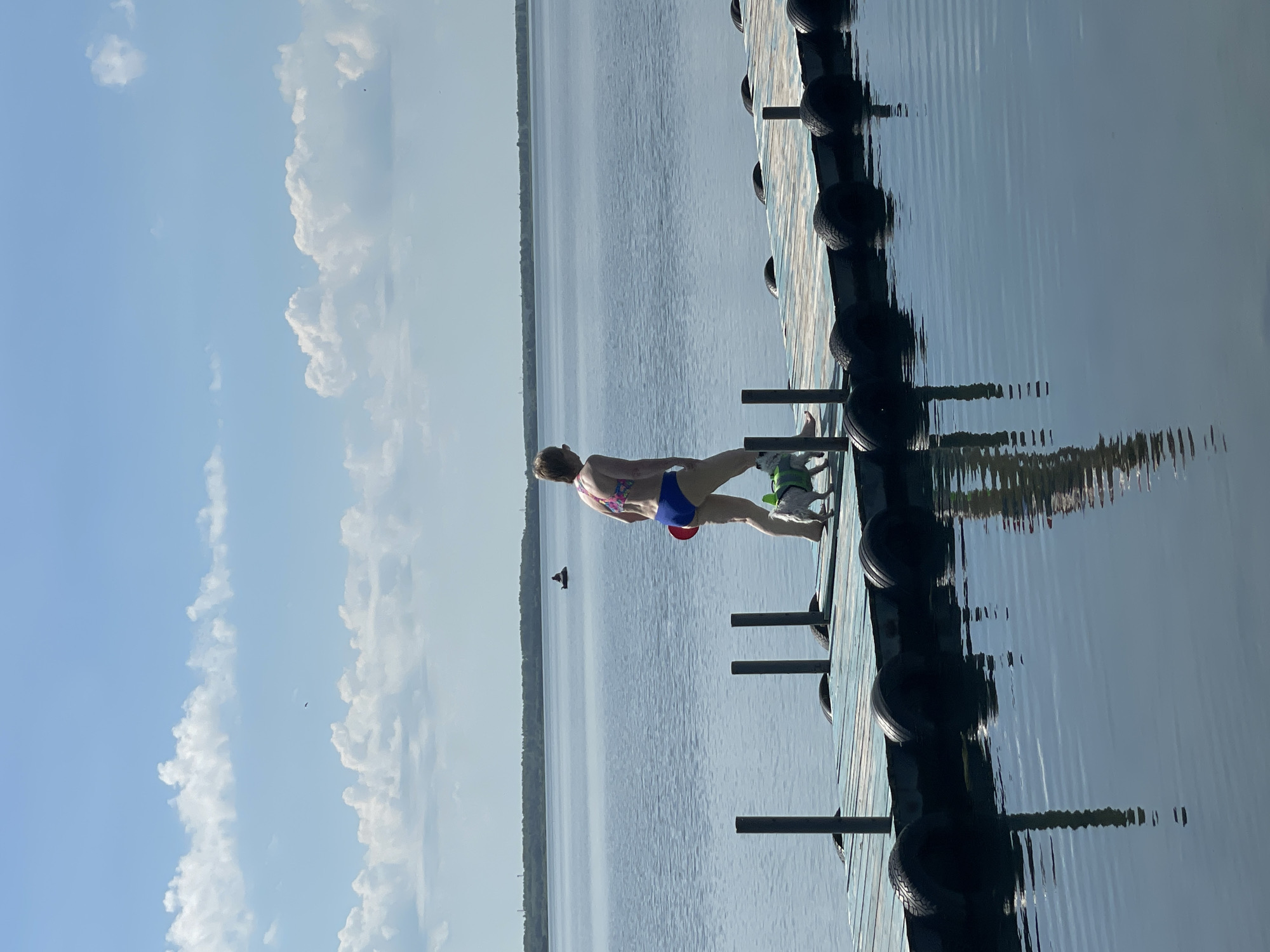 A woman in a blue bikini with a patterned top walks on a wooden dock at the edge of a calm lake. She is accompanied by a small white dog wearing a green life vest. The dog is walking in front of her. The dock has several vertical posts and is lined with tires. In the distance, across the lake, there is a tree line and a small boat with a person in it. The sky is blue with scattered white clouds, and the water reflects the sky and the dock.