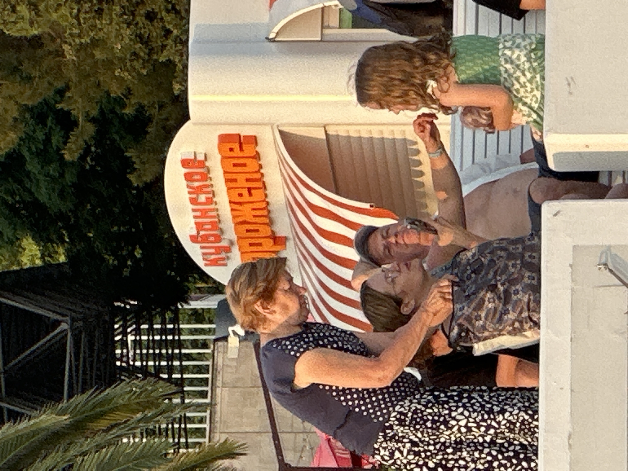 In the city of Sochi, Russia, an outdoor scene unfolds with a prominent sign featuring Cyrillic text in bright orange, reading "КУБАНСКОЕ МОРОЖЕНОЕ," which translates to "Kuban Ice Cream." The sign is set against a white building with arched awnings. In the foreground, several people are visible. An elderly woman, wearing a dark blue polka-dot top and a black and white patterned skirt, leans over to interact with a young girl. The girl, seated and wearing a green patterned dress, holds a doll. To the left of the girl, a man with a bare chest and a baseball cap is visible, and to the left of him, another woman with glasses is seated. Behind the group, a metal structure with diagonal supports and vertical bars is partially obscured by palm fronds. The overall lighting suggests it is late afternoon or early evening, with warm sunlight casting shadows.