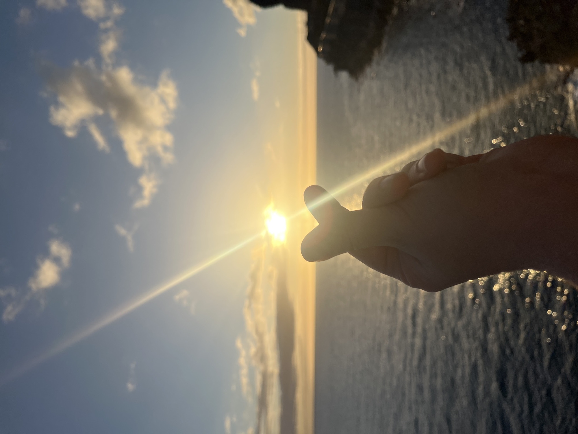 A hand forms a finger heart shape, partially obscuring the sun setting over the ocean. Some clouds are visible in the sky. The location is likely coastal, based on the presence of the ocean and possible cliffs in the background. The image is rotated 90 degrees. There is no evidence to support the location is the city of Puerto Rico, Spain.