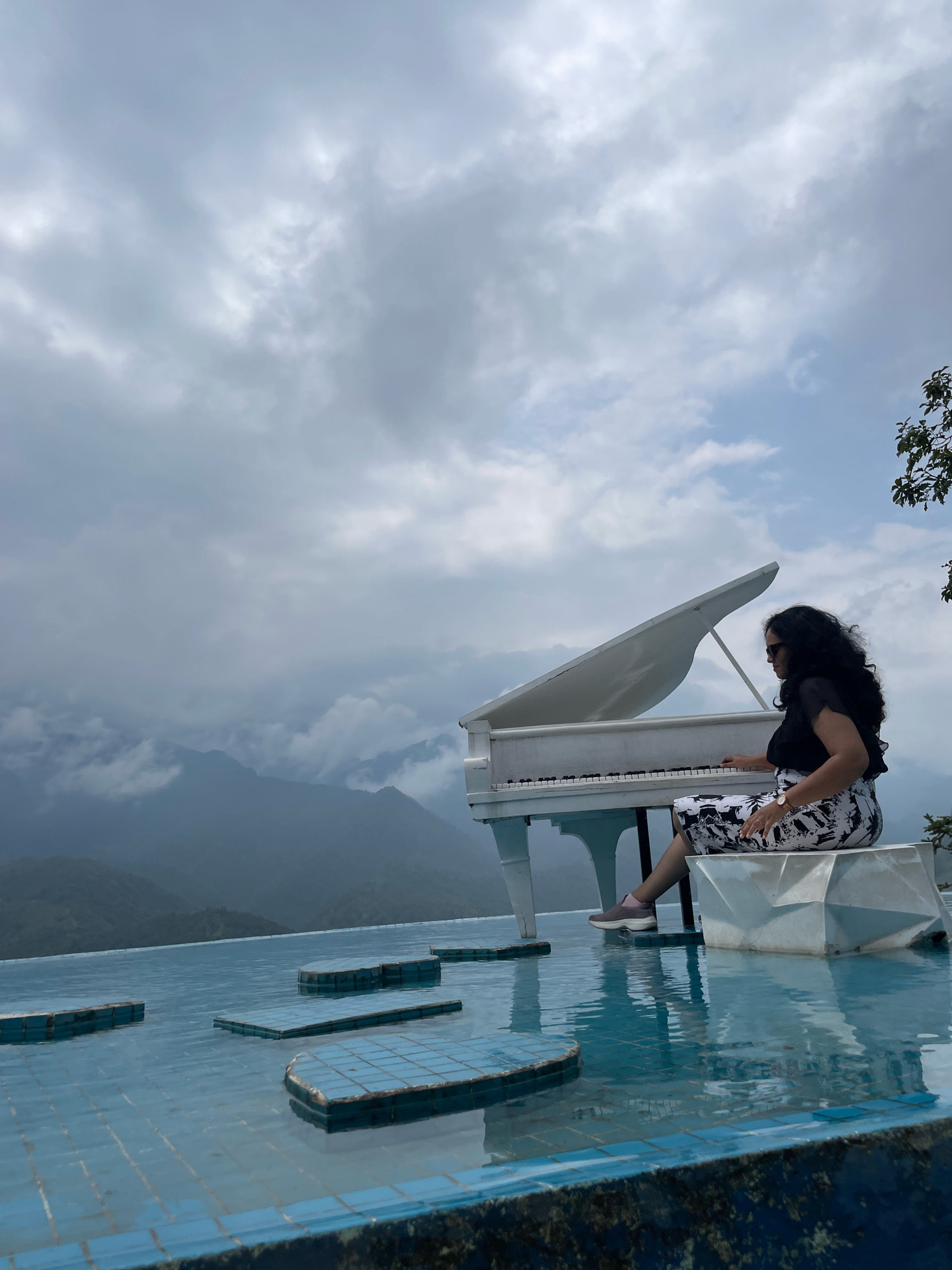 A woman with long, dark, curly hair and sunglasses is sitting on a white geometric stool in front of a white grand piano. She is wearing a black top and a black and white patterned skirt. Her right hand is on the piano keys, suggesting she is playing. The piano is positioned on the edge of an infinity pool, which is tiled with blue tiles. Several blue, tiled rectangular platforms are submerged in the pool, creating a visually interesting pathway. In the background, there are misty mountains and a cloudy sky. The overall scene suggests a scenic and artistic location, likely a hotel or resort in Sa Pa Ward, Vietnam, known for its mountainous landscape.