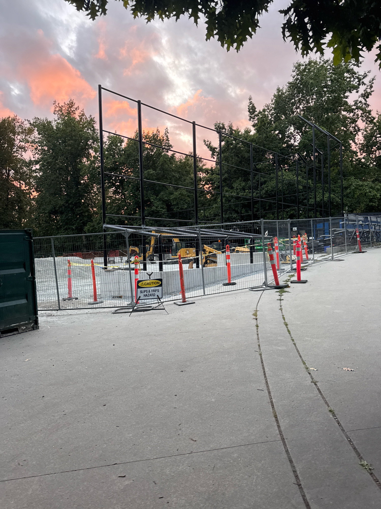 An outdoor construction site is shown in an image taken in Coquitlam, Canada. The sky above is filled with clouds tinged with pink and orange from sunset, contrasting with the dark silhouettes of trees. A tall black metal fence encloses an area under construction, where a concrete foundation is being laid. Orange traffic cones and a "CAUTION SLIPS & TRIPS HAZARD" sign are visible near the fence. Yellow construction equipment, including excavators, are present within the fenced area. A dark green shipping container is situated to the left of the frame. The ground in the foreground is paved with concrete, showing cracks and some sparse vegetation growing through them.