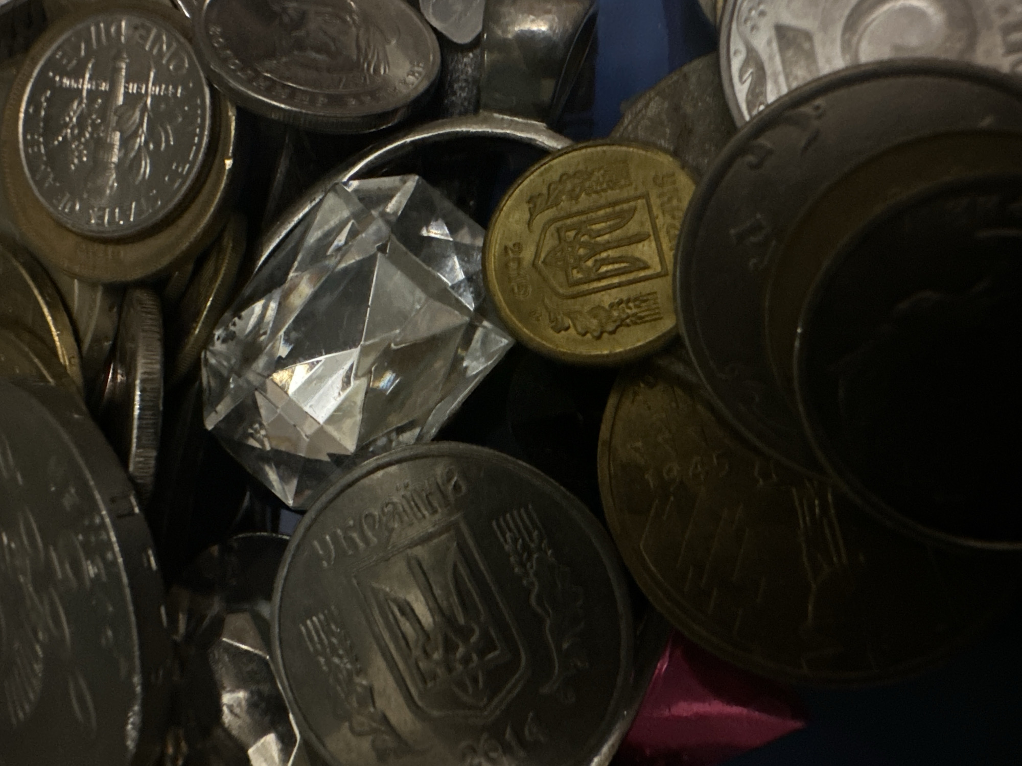 A close-up, overhead shot showcases a collection of coins and a large, faceted clear gem. The coins are of various sizes and metals, some appearing to be Ukrainian currency with their distinctive trident emblem and inscriptions in Ukrainian. One prominent coin, a golden hue, displays the year 2012. The gem, positioned centrally and angled, catches the light and exhibits sharp, geometric facets. The background is dark and indistinct, suggesting a shallow depth of field or a dimly lit environment. The composition is tight, with the objects filling the frame, creating a sense of abundance or a curated display. There is no evidence of human presence or a specific urban environment.