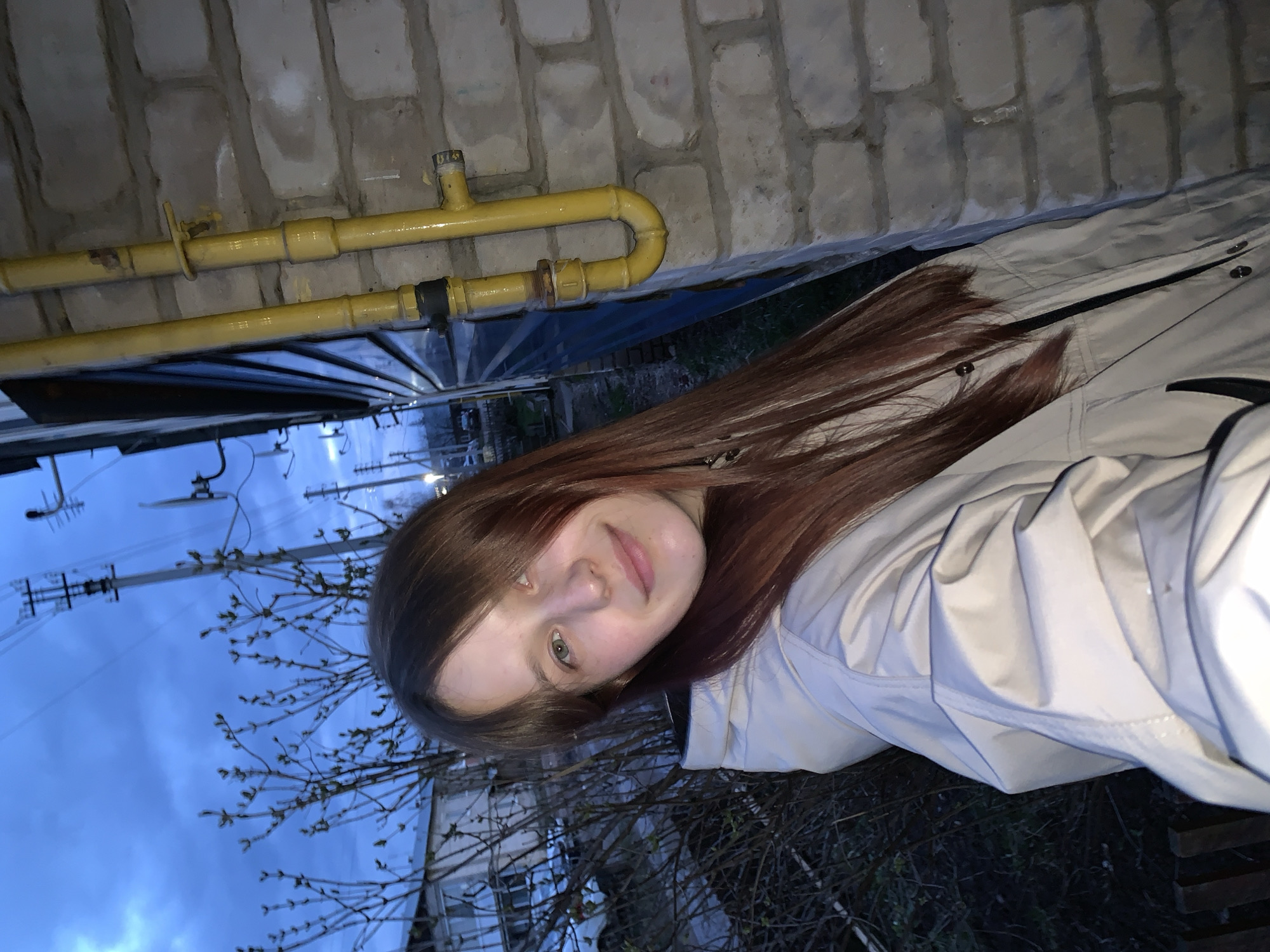 A young woman with long brown hair poses for a photo outside in Фоки, Russia. She is wearing a light-colored jacket. Behind her is a brick wall with yellow pipes running along it. In the lower portion of the image are bare tree branches against a twilight sky. Utility poles with wires are faintly visible in the background.