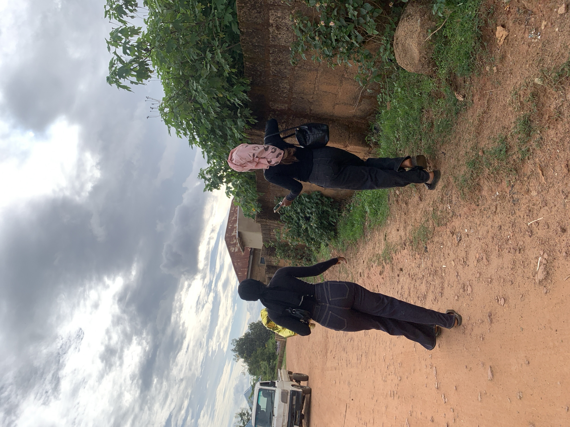 This image depicts a street scene in Jos, Nigeria, featuring two individuals observed from behind, walking away from the camera on an unpaved, reddish-brown dirt road. The individual on the left wears a black long-sleeved top, dark denim pants with visible stitching, and a black head covering, carrying a shiny gold, possibly inflatable, object in their left hand and a black shoulder bag. This individual wears dark, open-backed sandals. The individual on the right also wears a black long-sleeved top and dark denim pants, accessorized with a pink patterned head covering, a black handbag slung over their left shoulder, and a mobile phone held in their right hand. This individual also wears dark, open-backed sandals.
On the left side of the dirt road, a white compact pick-up truck is visible. In the mid-ground and background, residential structures with various roof materials, including one with a prominent reddish-brown corrugated roof, are present. The right side of the path is bordered by a rough, light brown block wall, with dense green foliage growing behind it and along the pathway. Several large rocks are visible near the wall. The sky is overcast with a mix of grey and white clouds, with brighter patches indicating obscured sunlight. The ground is characterized by packed dirt, showing tire tracks and footprints.