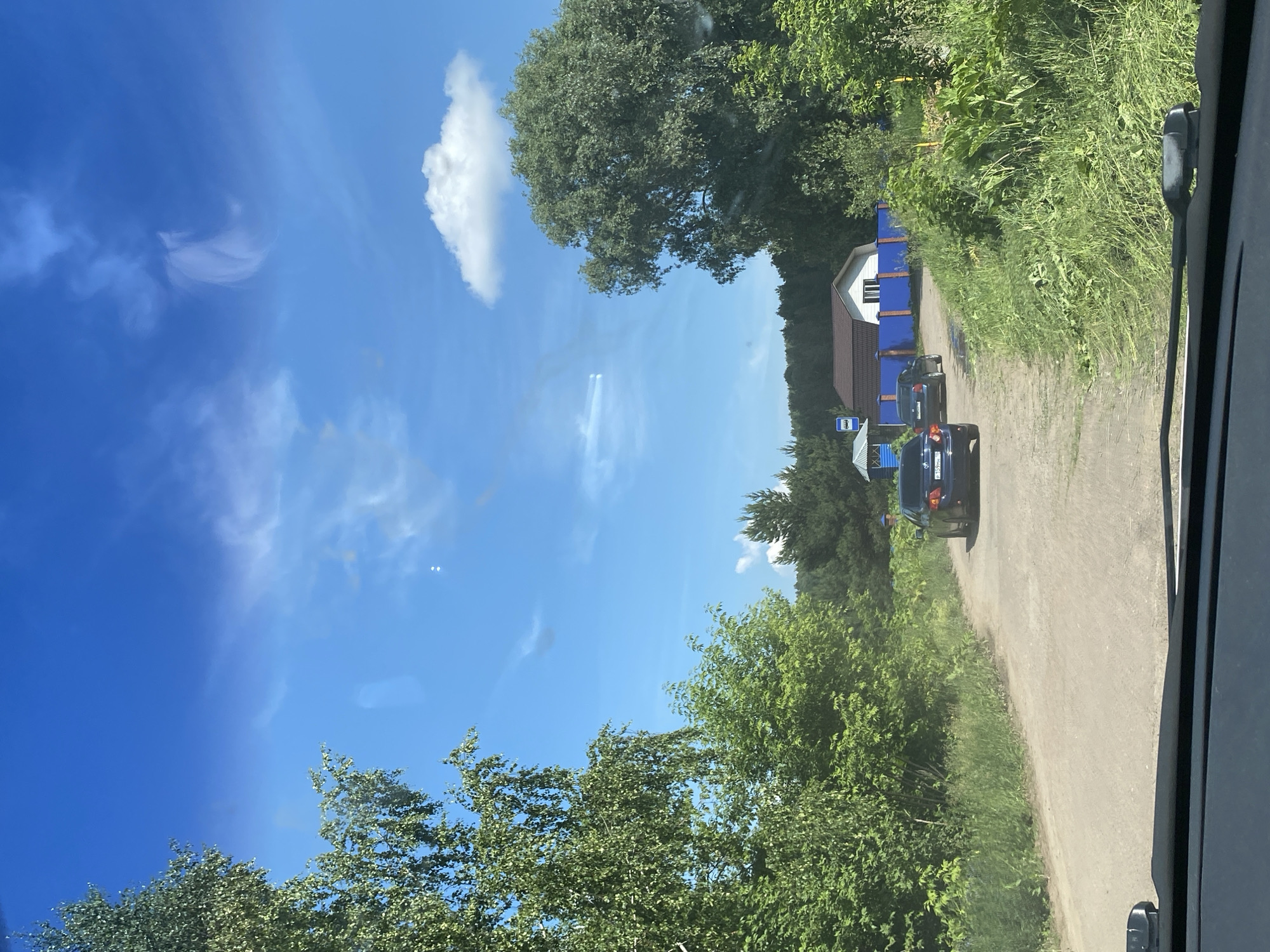 In the village of Evsino, Russia, a light-colored dirt road is visible, leading towards a cluster of houses. Two dark-colored sedans are driving on the road. The car in the foreground appears to be a dark blue sedan, and behind it is another dark sedan. Lush green trees and vegetation line both sides of the road. In the background, a white house with a brown roof is partially visible, bordered by a bright blue fence. A blue bus stop sign is also present. The sky above is a clear, bright blue with scattered white clouds. The image appears to be taken from inside a vehicle, looking out through the windshield.