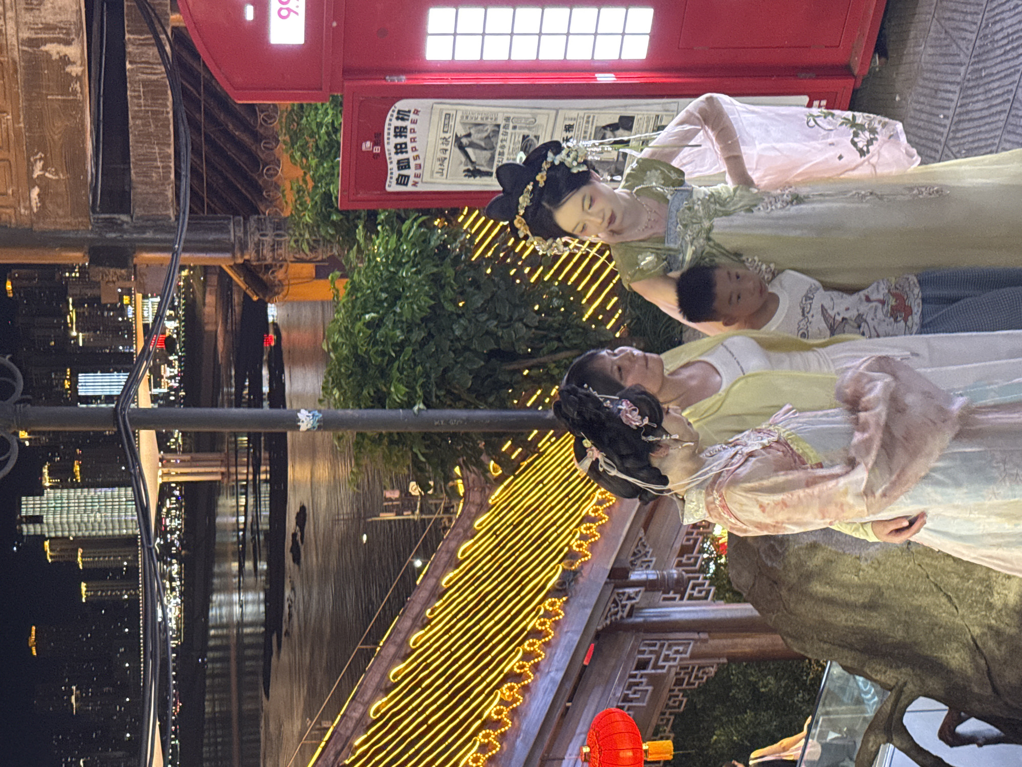 In Yuzhong District, China, three individuals, two women and one boy, are positioned in front of a vibrant red, phone booth-like structure. The woman on the right, adorned in a green, traditional Chinese dress with intricate floral embroidery and a floral headpiece, is looking to her left. To her left, a woman in a pale yellow jacket over a white top and a long white skirt stands, facing forward. A young boy, wearing a white t-shirt with a graphic print and checkered pants, stands between the two women, facing forward and slightly to his right. Behind them, a newspaper dispenser is visible, displaying Chinese text and images. The scene is illuminated by a backdrop of a city skyline at night, with illuminated buildings and a body of water reflecting the lights. A staircase adorned with bright yellow lights is to the left, and a red lantern hangs nearby, adding to the festive atmosphere.
