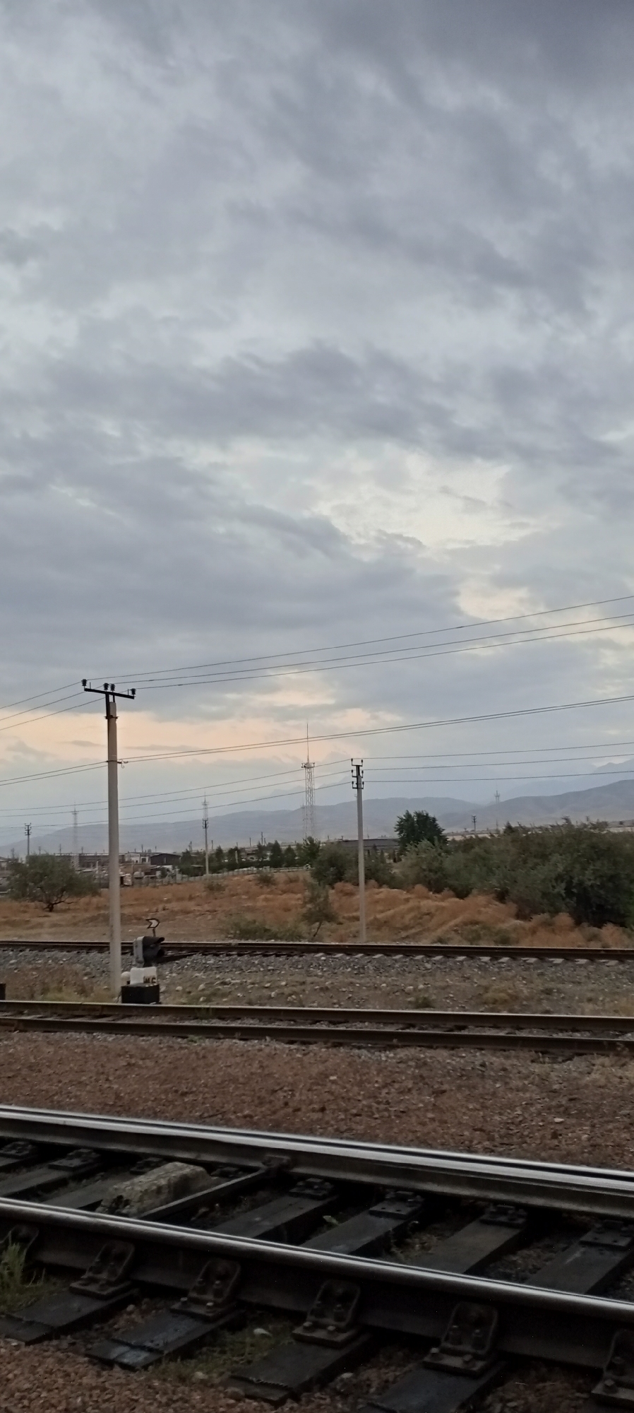 The image depicts a view of a railway line with overhead power lines and utility poles, likely in Sokuluk, Kyrgyzstan. The sky is overcast with a layer of gray clouds. In the middle ground, there are sparse dry grasses and some scattered trees, with a few buildings and a distant radio tower visible. The foreground features two sets of parallel railway tracks with gravel ballast. The overall scene is terrestrial and appears to be an outdoor landscape. There are no people or animals visible in the image.