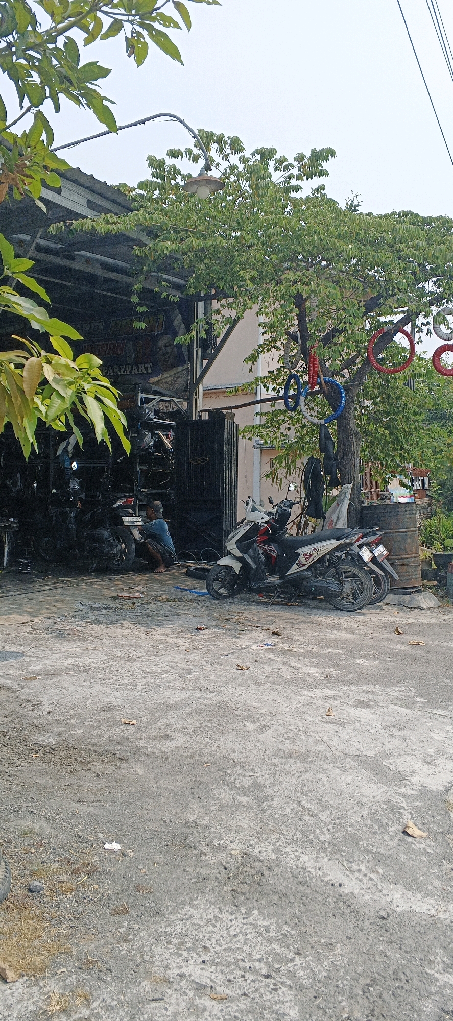 The image depicts an outdoor scene in front of a motorcycle repair shop in Sonoadi, Indonesia. A person in a blue t-shirt and dark shorts is crouched down, appearing to be working on a black motorcycle parked under a covered structure. The shop is filled with various motorcycles and spare parts. In front of the shop, two scooters are parked, one white and red, the other darker. A large tree with lush green leaves stands to the right, decorated with colorful rings, possibly made from recycled tires, hanging from its branches. Behind the tree, a large metal barrel is visible, and further back, a building with a light-colored facade. The ground is paved with rough concrete, with some scattered leaves and debris. The overall environment suggests a typical day at a local business in Indonesia.