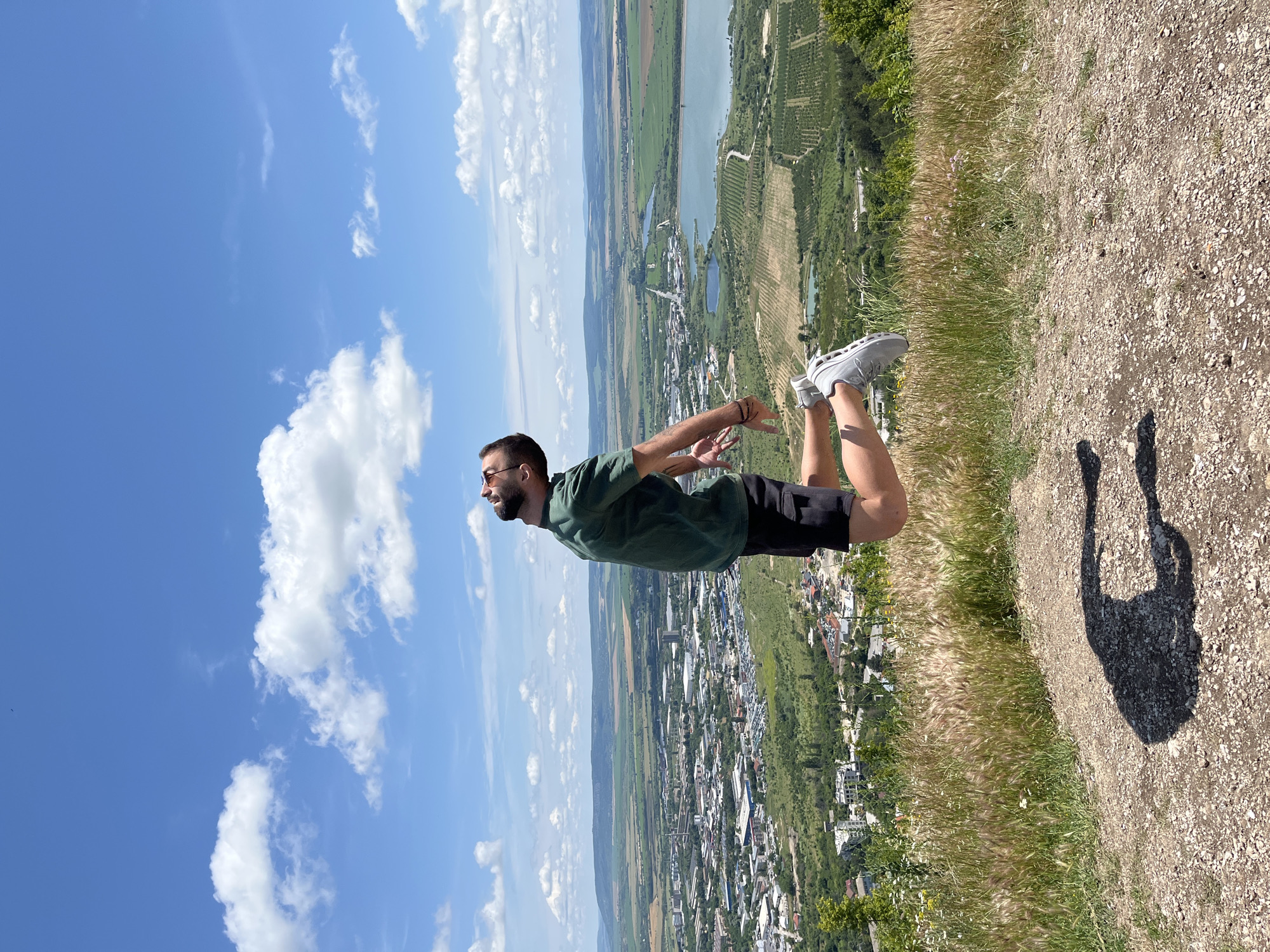 A man is captured mid-air in a jubilant jump against a backdrop of a vast urban landscape and rolling hills under a bright blue sky dotted with fluffy white clouds. The man, facing upwards with his head tilted back and eyes closed, is dressed in a dark green t-shirt, black shorts, and grey athletic shoes. His arms are bent at the elbows, and his legs are tucked towards his body, indicating the peak of his leap. The foreground is a rough, gravelly path bordered by dry, wispy grass and a few scattered wildflowers. His shadow is cast on the ground below, mirroring his jumping pose.
The sprawling city of Shumen, Bulgaria, unfolds in the mid-ground, featuring a dense collection of buildings, industrial structures, and residential areas. A prominent body of water, likely a reservoir or lake, is visible to the right of the city, surrounded by agricultural fields with distinct patterns of cultivation. The landscape extends into the distance with layers of green hills under the expansive sky. The overall scene conveys a sense of freedom and exhilaration, with the man's dynamic pose contrasting with the static panorama of the city and nature.