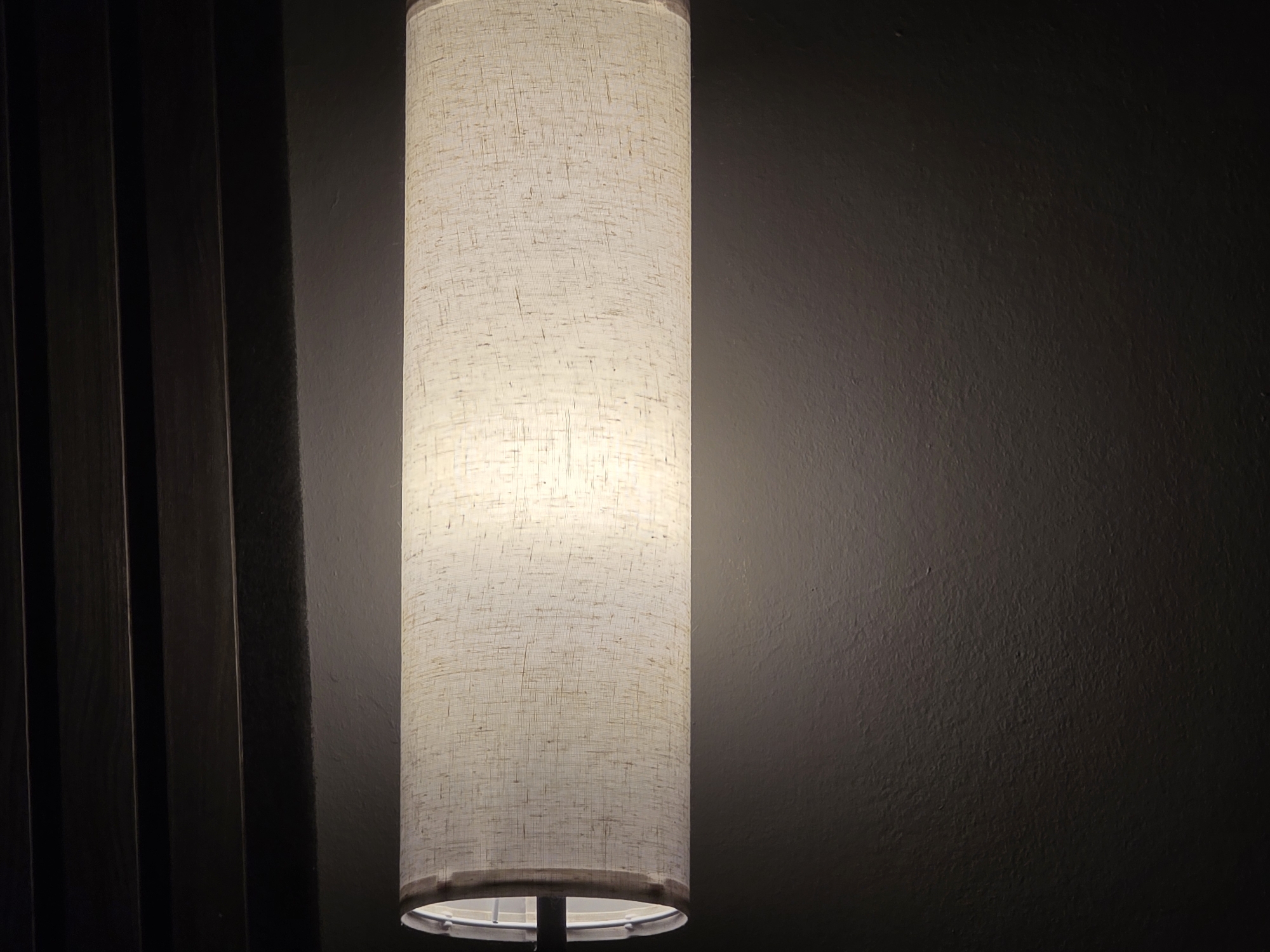A tall, cylindrical lampshade made of a light-colored, textured fabric is illuminated from within, casting a warm glow. The lampshade is positioned vertically, with the light source visible at the bottom. To the left of the lampshade, there are dark, vertical wooden slats, creating a textured background. To the right, the wall is a dark, textured grey. The scene appears to be an indoor setting, possibly a room in a hotel or apartment in Voluntari, Romania. There are no people or any discernible actions taking place. The overall impression is one of low lighting and a calm atmosphere.