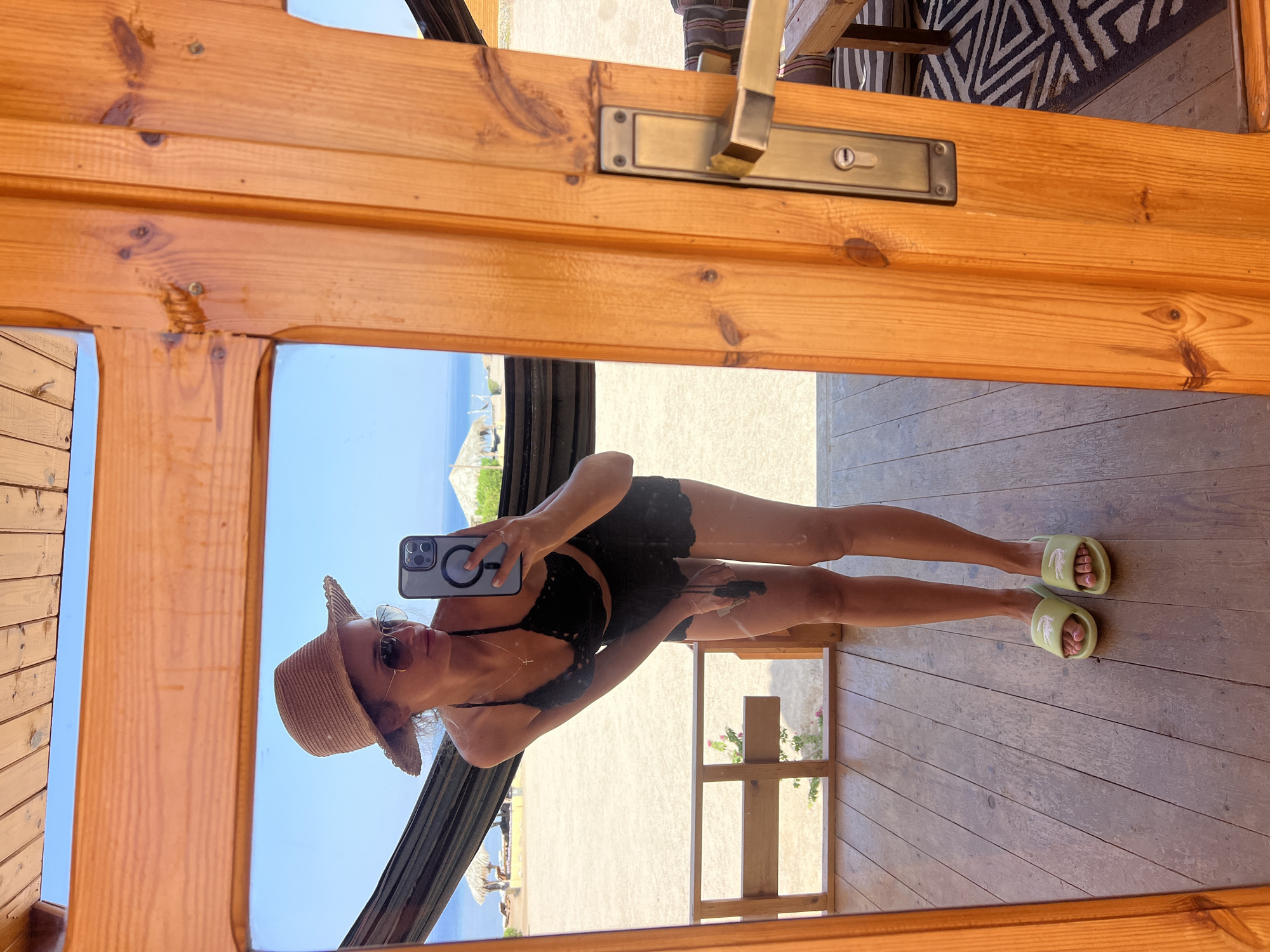 A woman stands on a wooden deck, taking a mirror selfie. She is wearing a straw hat, sunglasses, a black lace bikini top and shorts, and light green slides. In her hand, she holds a smartphone, which she uses to capture her reflection. The background reveals a sandy beach with a hammock, palm trees, and thatched-roof structures, with the ocean and clear blue sky visible beyond. The scene is likely in Nueiba, Egypt, a coastal city known for its natural beauty and laid-back atmosphere. The wooden structure she is in appears to be an open-air room or porch, with a wooden door frame and a metal door handle visible on the right side of the image.