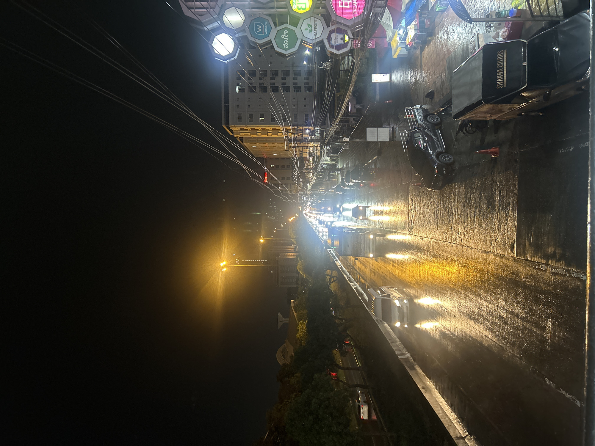 A street-level view captures a wet highway in Khlong Luang Town Municipality, Thailand, at night. Vehicles, including a white van and a truck with a trailer, travel on the glistening, rain-soaked road, reflecting the bright yellow streetlights. On the right side of the road, an apartment building is illuminated, with power lines crisscrossing the sky. Several shops and market stalls are visible, with one van bearing the signage "SIVANNA COLORS." A black pickup truck is parked near the roadside, and various traffic cones are present. The sky is dark, indicating nighttime.