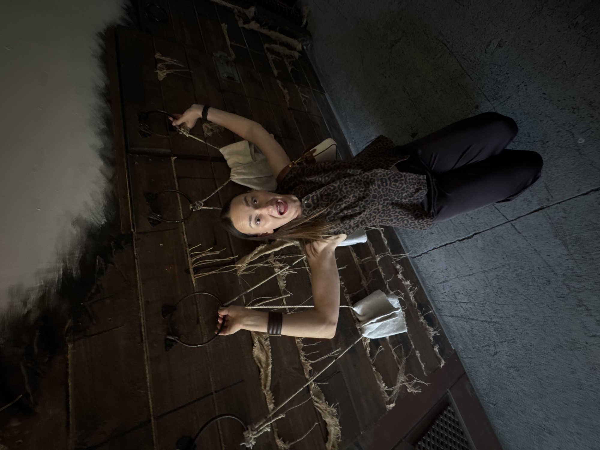 A woman is kneeling on a concrete floor, her arms raised and gripping rings attached to a wooden wall. The wall is covered in burlap and ropes, and appears to be part of a theatrical set or exhibit. The woman is wearing a leopard-print top and dark pants, and her mouth is open in an exaggerated expression of surprise or distress. The setting evokes a dungeon or prison cell. The location is identified as Sochi, Russia, though this specific context is not visually apparent from the image itself.