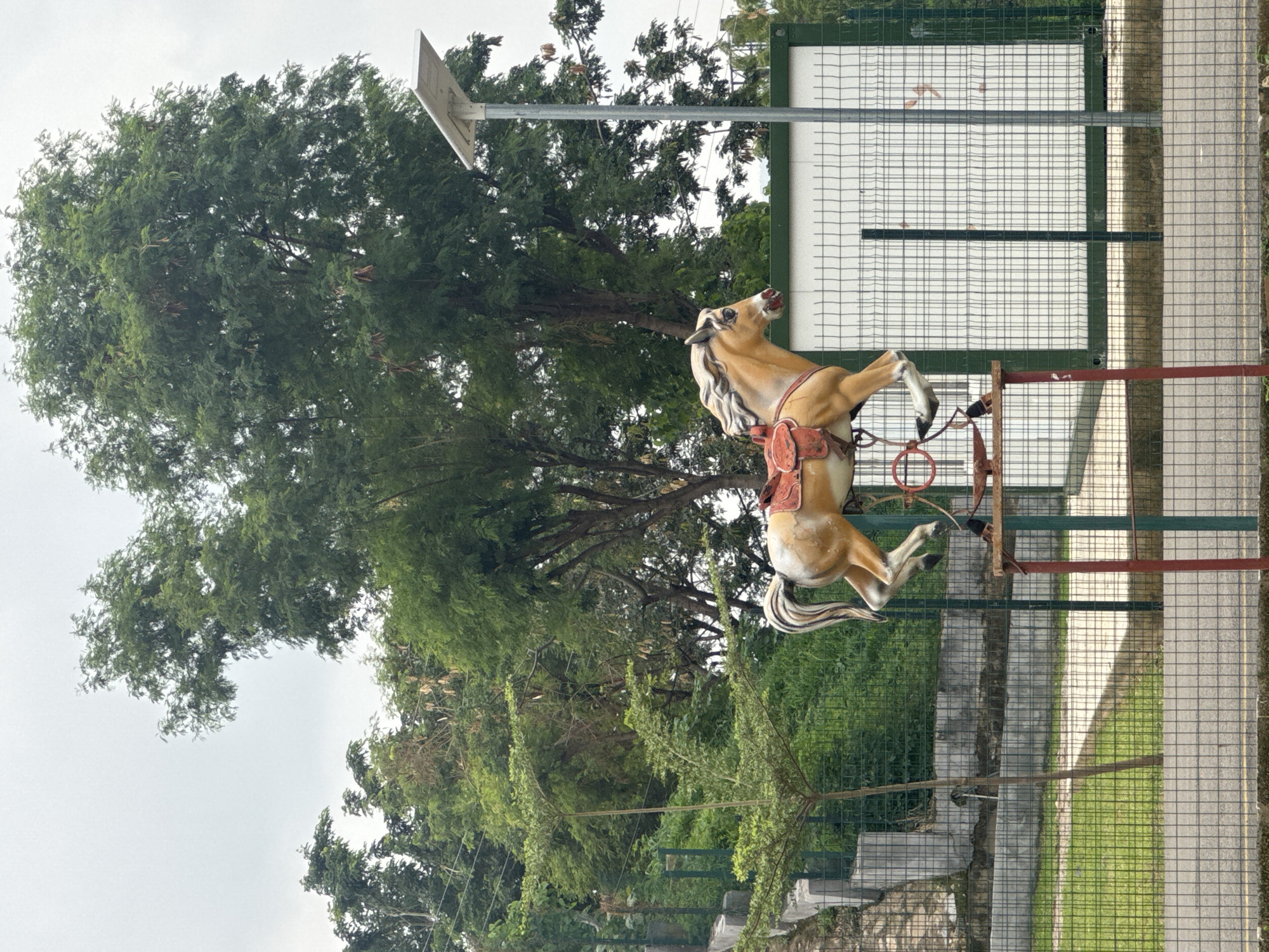The image depicts a stationary carousel horse in an outdoor setting. The horse is light brown with a white mane and tail, equipped with a saddle. It is mounted on a red and grey metal frame. Behind the horse is a green metal fence and a paved path. The background features a large tree with dense foliage and a utility pole with a solar panel. The location is Abuja, Nigeria.