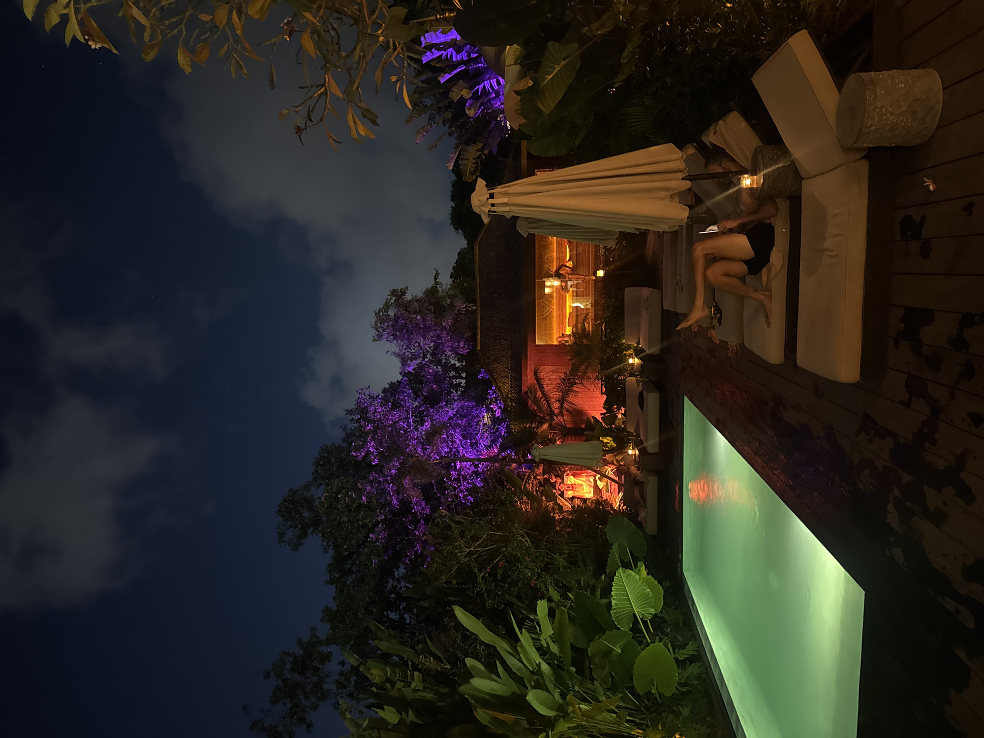 A nighttime photograph taken in Mengwi, Indonesia, shows an outdoor lounge area. A person is reclined on a white lounge chair beside a lit rectangular pool. The person appears to be using a tablet or phone. An open white umbrella partially covers the lounge area. Trees and foliage surround the area, some lit with purple and orange lights. The sky is visible with scattered clouds.