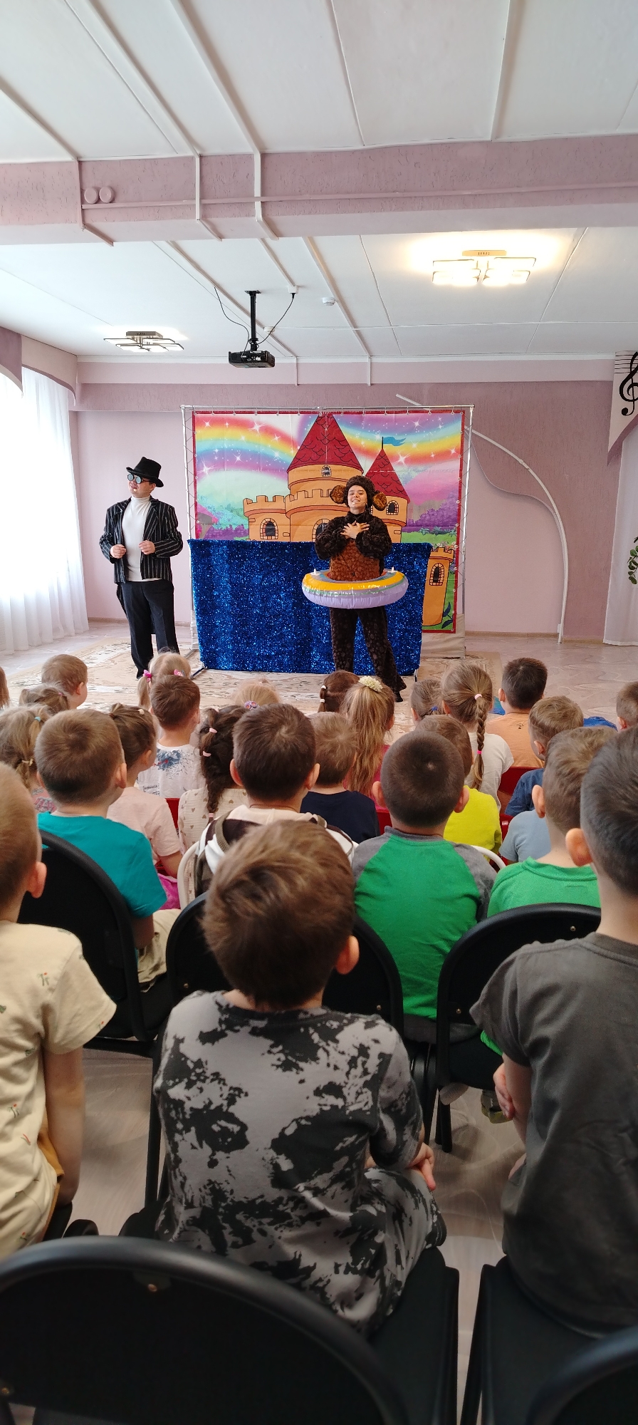 The image shows a room full of children sitting in chairs, facing a stage. On the stage are two people in costume. One is wearing a black suit with white stripes, a white turtleneck, a black hat, and sunglasses. The other is wearing a brown animal costume with an inflatable ring around their waist. Behind them is a backdrop depicting a castle and a rainbow. The room has pink walls and a white ceiling. A projector is hanging from the ceiling. The image was taken in Chaikovsky, Russia.