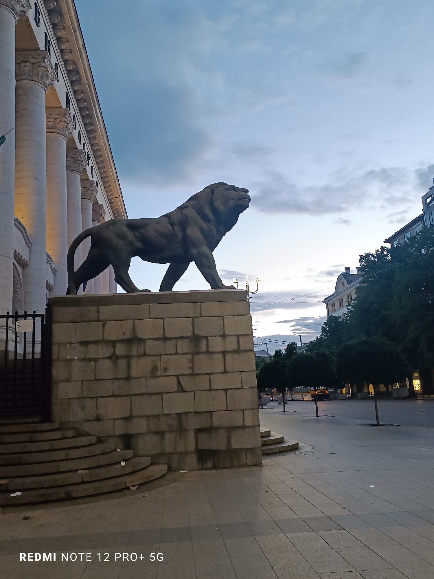 The image shows a lion statue at the Palace of Justice in Sofia, Bulgaria, viewed from a low angle. The statue is dark gray, positioned atop a multi-tiered stone base constructed of large blocks. Part of the building's facade is visible to the left, with prominent white columns. A dark iron gate sits adjacent to the statue. To the right, a street scene includes trees, buildings, and streetlights. The sky is overcast, with varying shades of gray and hints of light. The ground is paved with large rectangular slabs. A "Redmi Note 12 Pro+ 5G" watermark is visible at the bottom.