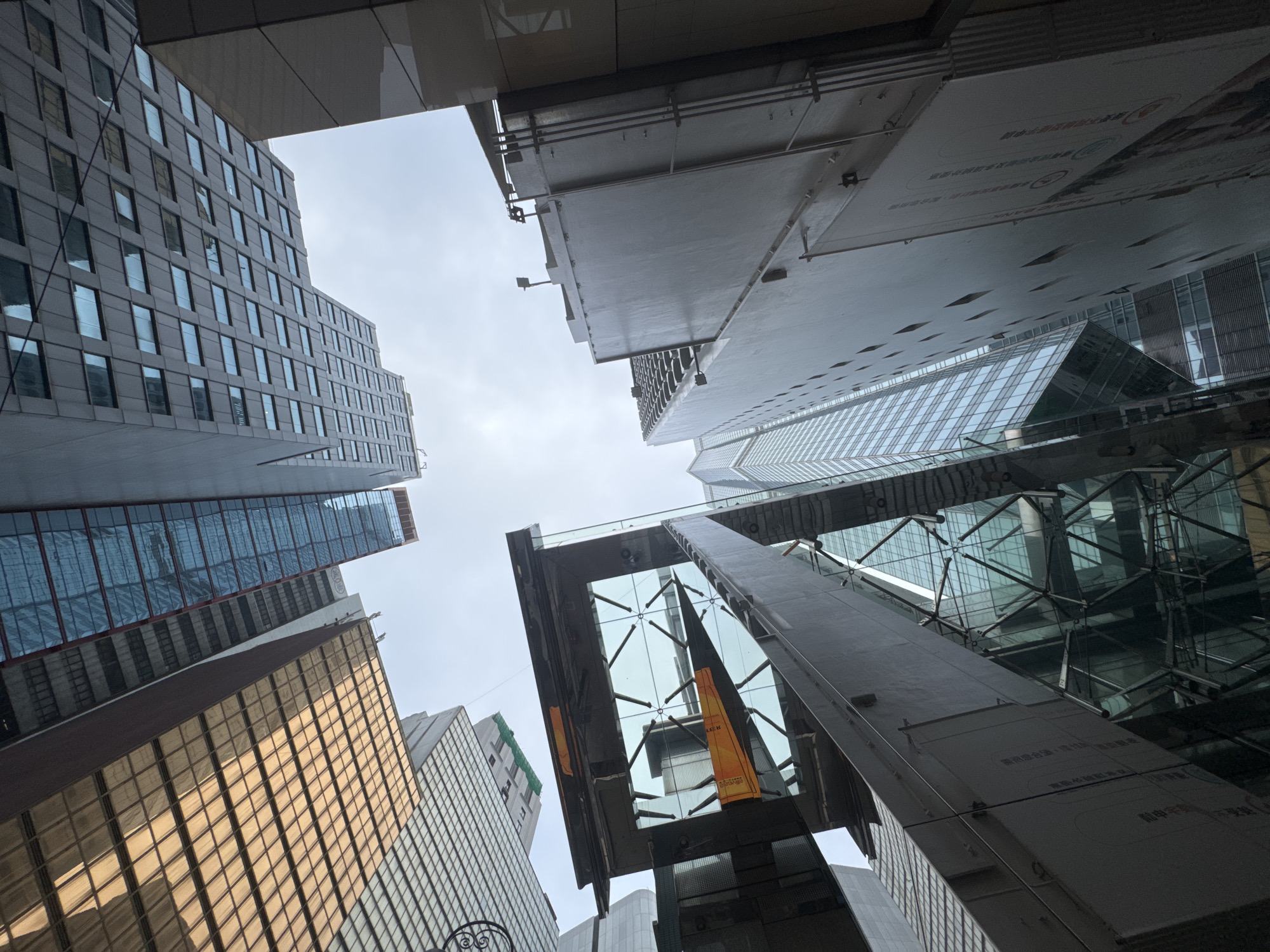 The image depicts a low-angle view of several skyscrapers in Hong Kong, China. The architectural styles are modern, with glass and steel facades dominating. The buildings vary in color and texture, with some reflecting the sunlight and sky. In the center of the image, a unique building structure stands out. The environment shows a slightly overcast sky. There are no discernible people or actions.