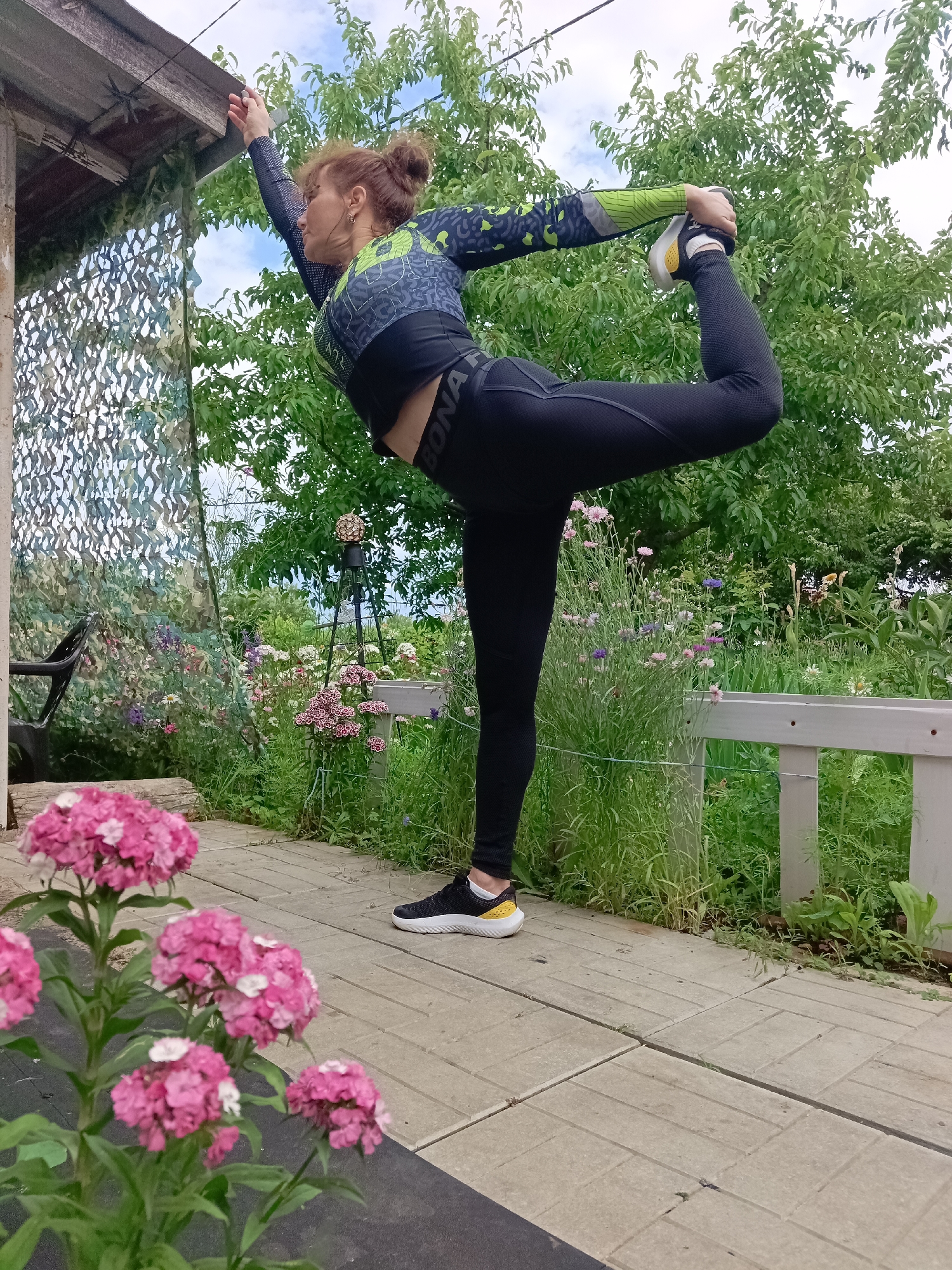 A woman with red hair tied in a bun, wearing a long-sleeved black and green athletic top and black leggings, performs a yoga pose on a paved outdoor patio. She stands on her left leg, with her right leg bent at the knee and her right foot held in her right hand. Her left arm is extended upwards. In the background, there is lush green foliage, flowering plants in shades of pink and purple, a wooden structure with camouflage netting, and a light-colored fence. The sky is partly cloudy. The scene appears to be set in a garden or backyard. The location is identified as Vyazovka, Russia.