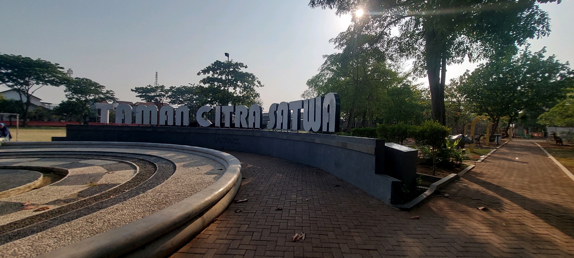 The image shows a large, white sign with the words "TAMAN CITRA SATWA" in large, 3D letters. The sign is situated in front of a dark gray, curved wall. The ground in front of the wall is paved with reddish-brown bricks arranged in a herringbone pattern. To the left of the sign and wall is a curved, tiered structure with a textured surface, possibly a fountain or decorative seating area. Beyond the sign and wall, a park-like setting is visible with lush green trees, bushes, and a pathway. In the background, there are buildings, a communication tower, and a clear sky with sunlight filtering through the trees, creating long shadows. The overall scene suggests an outdoor public space, likely a park or recreational area. The phrase "TAMAN CITRA SATWA" translates to "Satwa Image Park" or "Satwa Nature Park," and the presence of animal statues in the background (a rhino and a deer-like creature, and possibly a bird or dinosaur statue further down the path) reinforces the theme of a nature or animal-themed park. This specific sign is located in Taman Citra Satwa, a zoo and recreational park in Semarang, Indonesia.