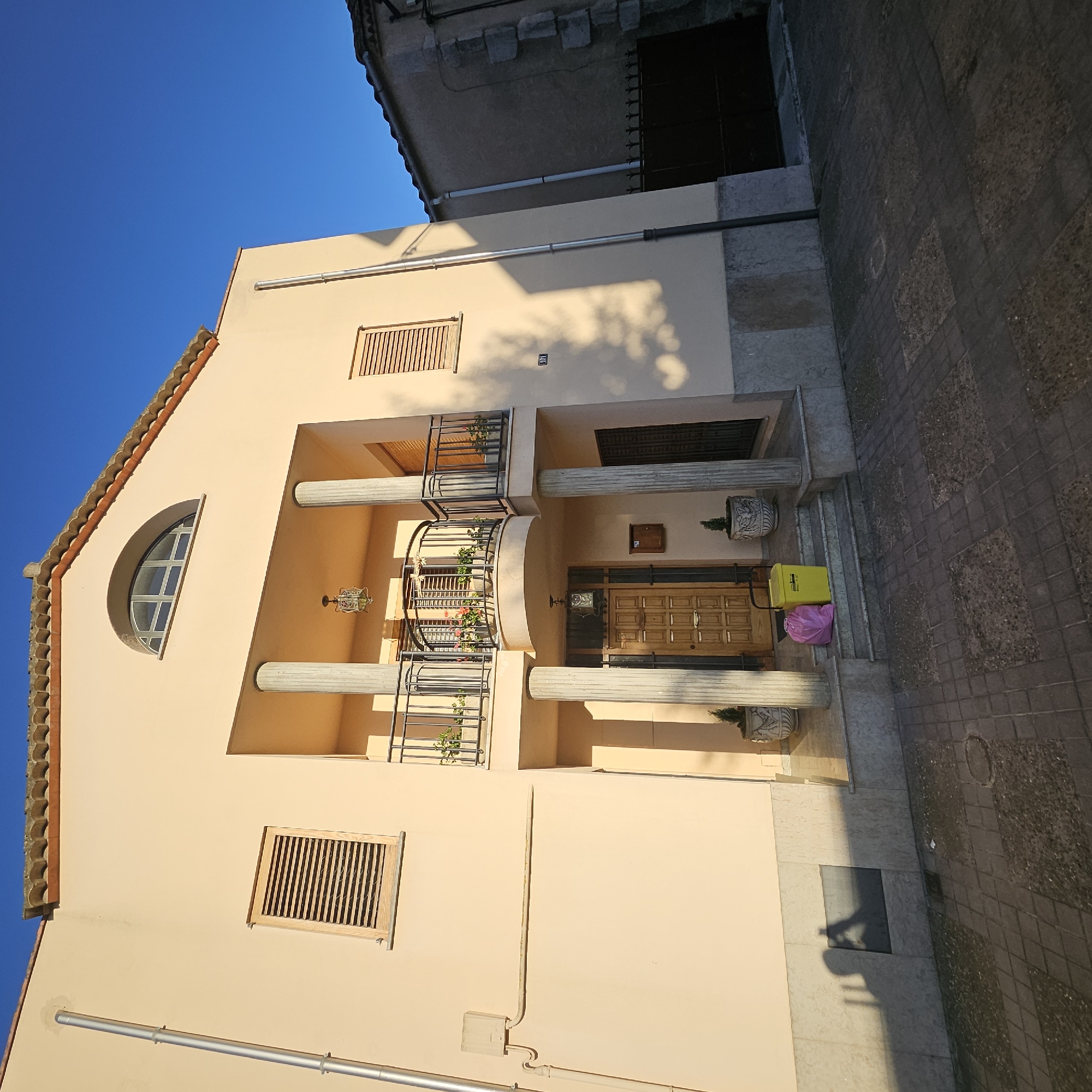 A residential building, likely a house, in Vilablareix, Spain, is shown in this exterior, low-angle, wide shot. The building has a pale orange facade, a terracotta-tiled roof, and an arched window at the top. A balcony with black metal railings is supported by two large columns. The entrance to the building features a wooden door and a small set of stairs leading up to it. There are potted plants on the steps and on either side of the entrance. A yellow recycling bin and a pink trash bag are placed on the steps. A shadow of a person holding a camera is cast on the left side of the building. The sidewalk in front of the building is paved with square tiles. To the right of the building, another residential structure with a grey facade and stone accents is visible, along with a dark metal gate. The sky is a clear, bright blue. The scene is illuminated by sunlight, casting shadows and highlighting the architectural features of the buildings.