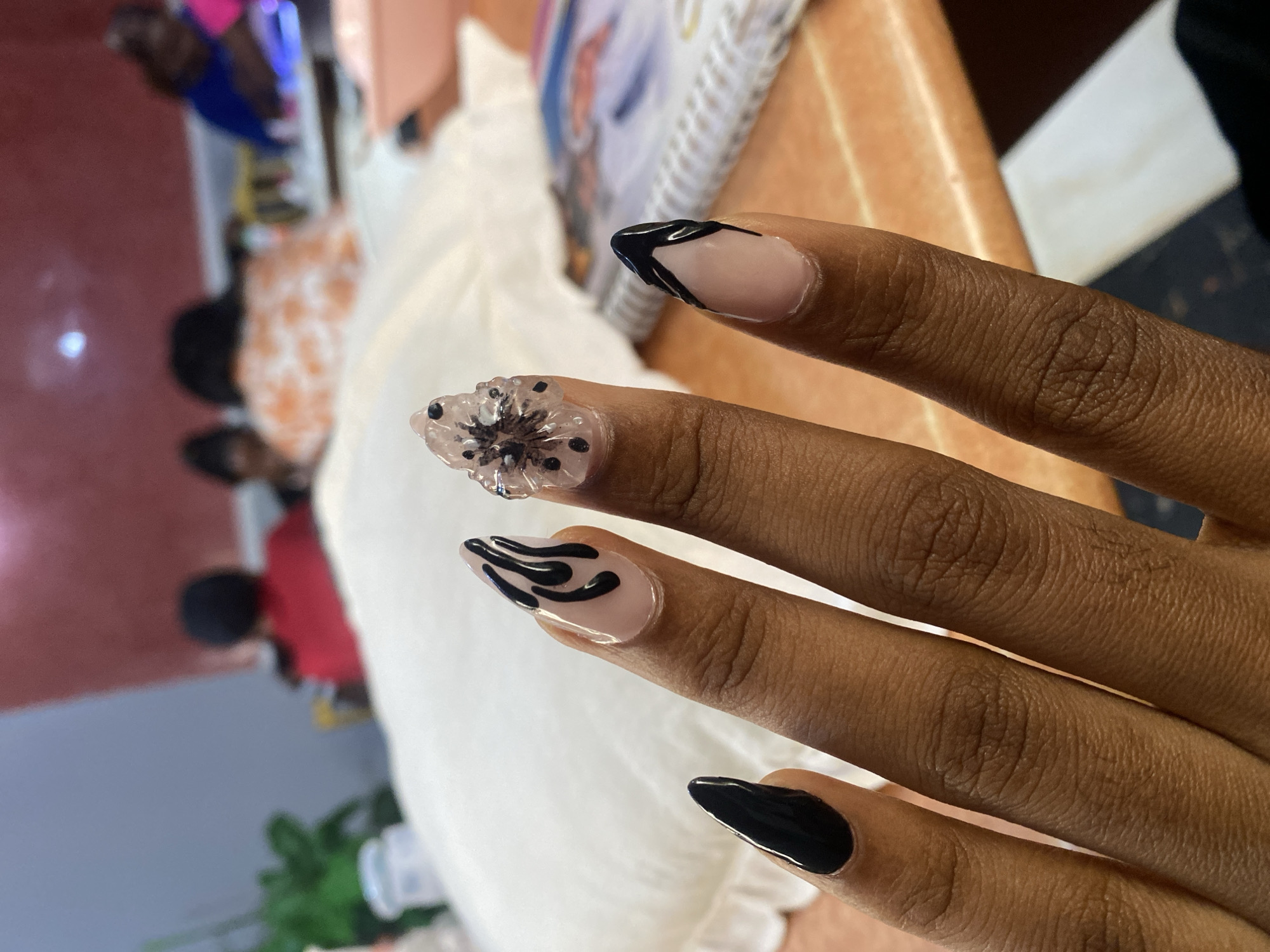 The image shows a close-up of a person's hands with elaborate nail art, likely taken in a nail salon. The nails are long and stiletto-shaped, with various designs including black flames, a clear overlay with flower detailing and black dots, and a French tip with black accents. In the blurred background, several people are visible, some wearing colorful clothing, suggesting a public or communal setting. The overall environment appears to be indoors, with a table in the foreground and what looks like a white tablecloth or covering. The lighting suggests it could be daytime or under artificial light. The location in Jos, Nigeria, cannot be determined from the visual information provided.