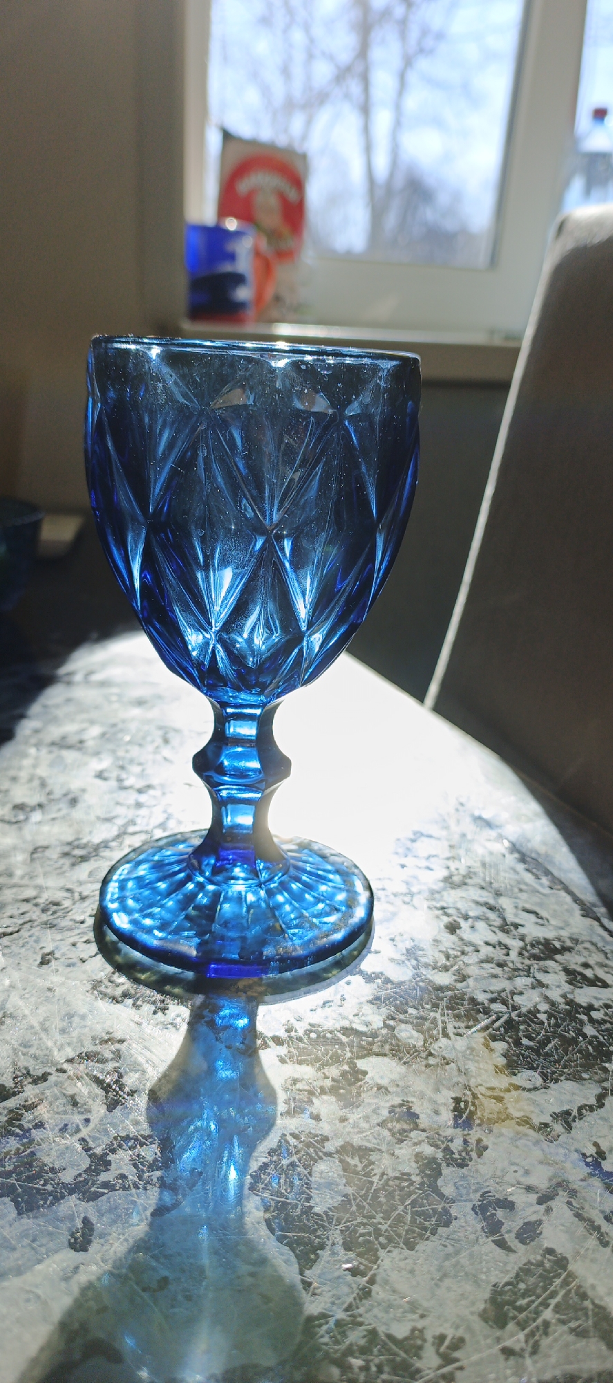 A vibrant, blue, cut-glass goblet sits on a surface resembling marble. The sunlight streams in from a window behind the goblet, creating a bright shadow of the glass on the tabletop. The shadow mimics the shape of the glass itself, casting a distorted blue hue onto the surface. In the background, through the window, blurry glimpses of a building, possibly with advertisements, and bare trees suggest an urban setting. A can or bottle with bright red label is visible. The environment suggests the location could be Chaikovsky, Russia. A partial view of a chair back is visible on the right side of the image.