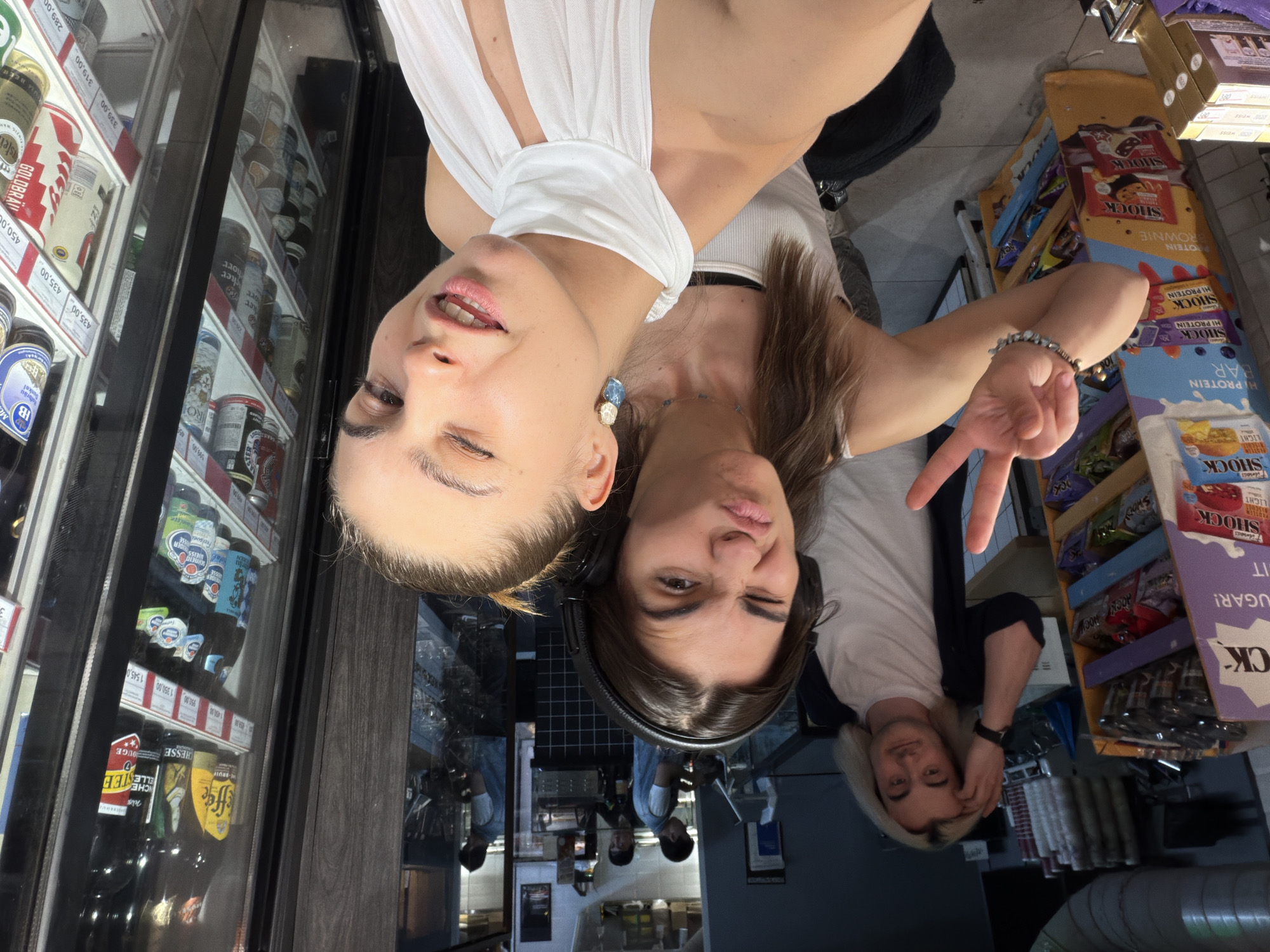Three young adults, two women and one man, are taking a selfie in what appears to be a convenience store or small grocery store. The woman in the foreground is wearing a white halter top and a large blue and white earring, and is winking at the camera. The woman behind her is wearing headphones and making a funny face. The man behind them is touching his hair and looking at the camera. There are shelves of snacks and drinks in the background, as well as a refrigerated display case filled with bottles of beer. The scene is lit by fluorescent lighting. The text on the snack wrappers indicates that this is a Russian store, as does the text on the price tags in the beer display. The location is identified as Moscow, Russia.