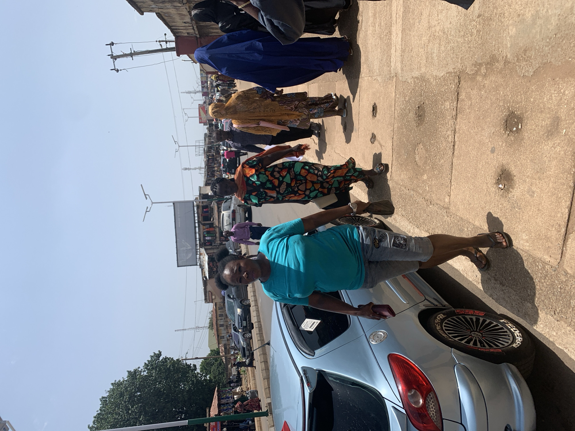 In the city of Jos, Nigeria, on a sunny day, a woman in a turquoise t-shirt and denim shorts walks on a sidewalk next to a light blue car. She carries a dark red purse in her left hand. Behind her, a group of people are walking along the sidewalk. One woman wears a colorful, patterned dress, another is draped in a bright yellow fabric, and a third is covered in a dark blue abaya. Further down the street, traffic is visible, including cars and a bus, with buildings and shops lining the road. The sidewalk itself is made of concrete slabs, some of which appear worn and have holes. The scene depicts a typical urban street in Jos, with bustling pedestrian and vehicle activity under a clear sky.