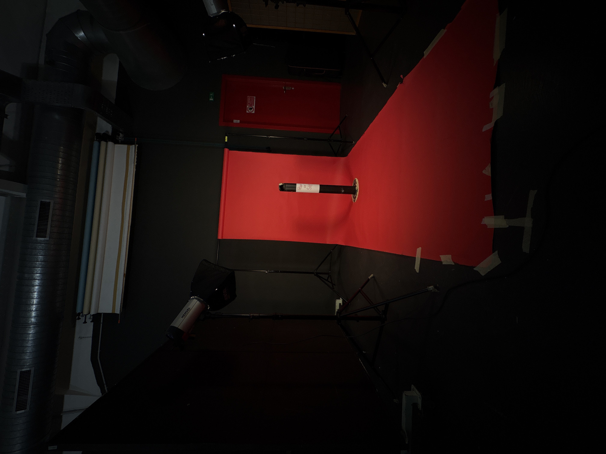 This image depicts the interior of what appears to be a photography or video studio, potentially in Milan, Italy, as indicated by the location context. The primary focus is a vivid red seamless backdrop, curving from the floor up to the wall, creating a continuous surface for shooting. White tape is visible along the edges, securing the backdrop to the dark walls.
In the center of the red backdrop, a black and white cylindrical object, resembling a stand or pipe, stands vertically. A white label on this object partially reads "The red..." with the subsequent line appearing as "...of SHE" or similar text.
Studio lighting equipment is visible, notably a silver Hensel strobe light with a black softbox or reflector attached, positioned on a stand to the left. Another large black softbox or flag is placed on the floor in the immediate foreground. Various stands and supports are scattered around the setup.
In the background, to the left, large corrugated metal pipes are prominent along the walls and ceiling. Below these pipes, several rolls of seamless paper in different colors (including white, beige, and light blue) are stored upright. To the upper right, a red utility door with a white warning sign, featuring a red circle and diagonal line (indicating 'no entry' or a similar restriction), is visible.
The scene is dimly lit overall, typical of a controlled studio environment where specific lighting is used for the subject. No individuals are present, suggesting the image captures the studio in preparation for a shoot or between sessions.