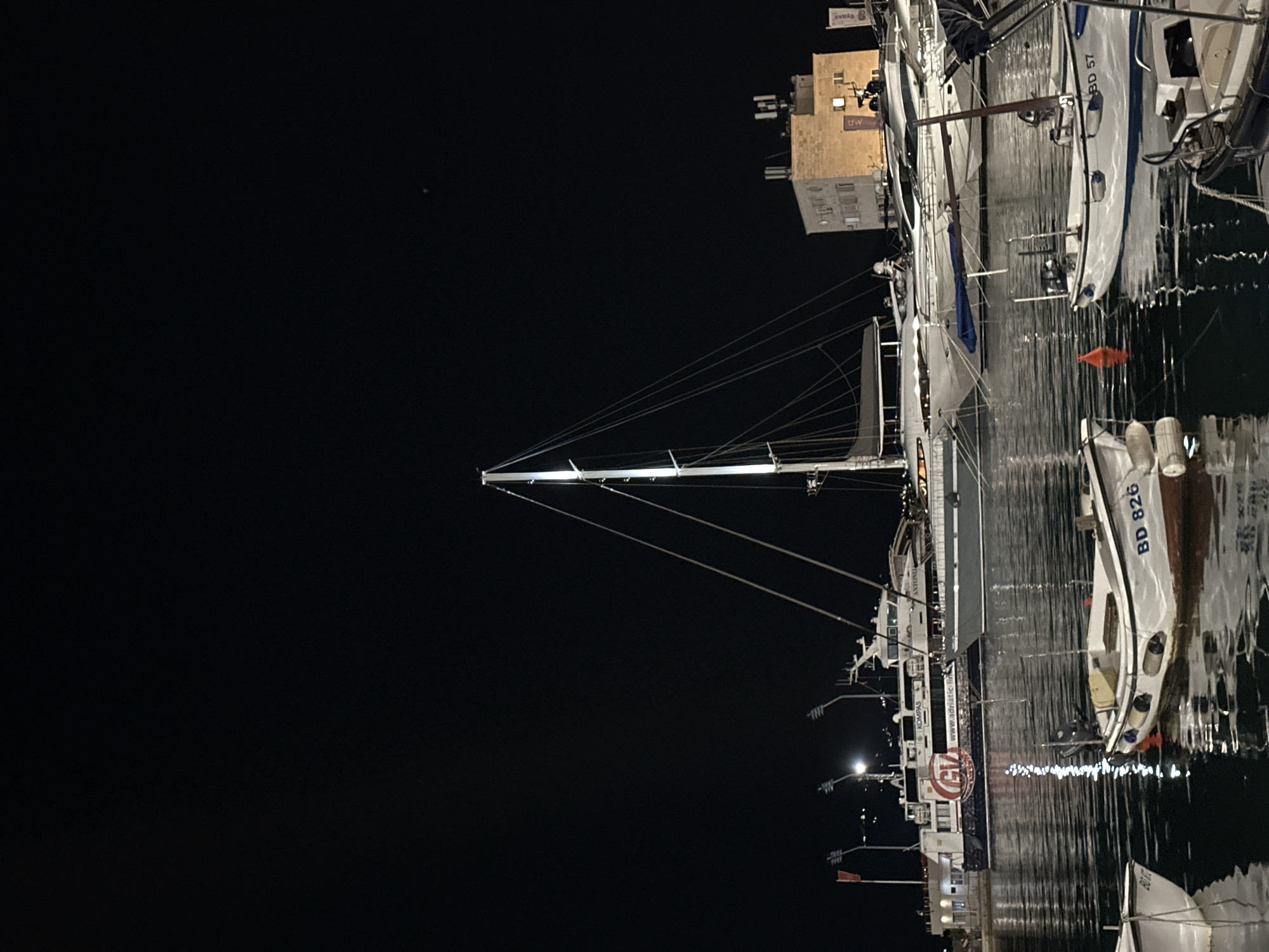 This is a nighttime photograph of a harbor in Budva, Montenegro, with several boats and a large ferry docked. In the foreground, two small white boats are visible, one with the registration "BD 826". In the middle ground, a large ferry with "GV" and "Adriatic Lines" branding is docked. Behind the ferry, a tall sailboat mast is prominently featured, with its rigging illuminated. Further in the background, a building with several windows and antennae is visible, with lights on inside. The water is calm and reflects the lights of the boats and the buildings. The sky is dark and clear, with a few distant lights visible.