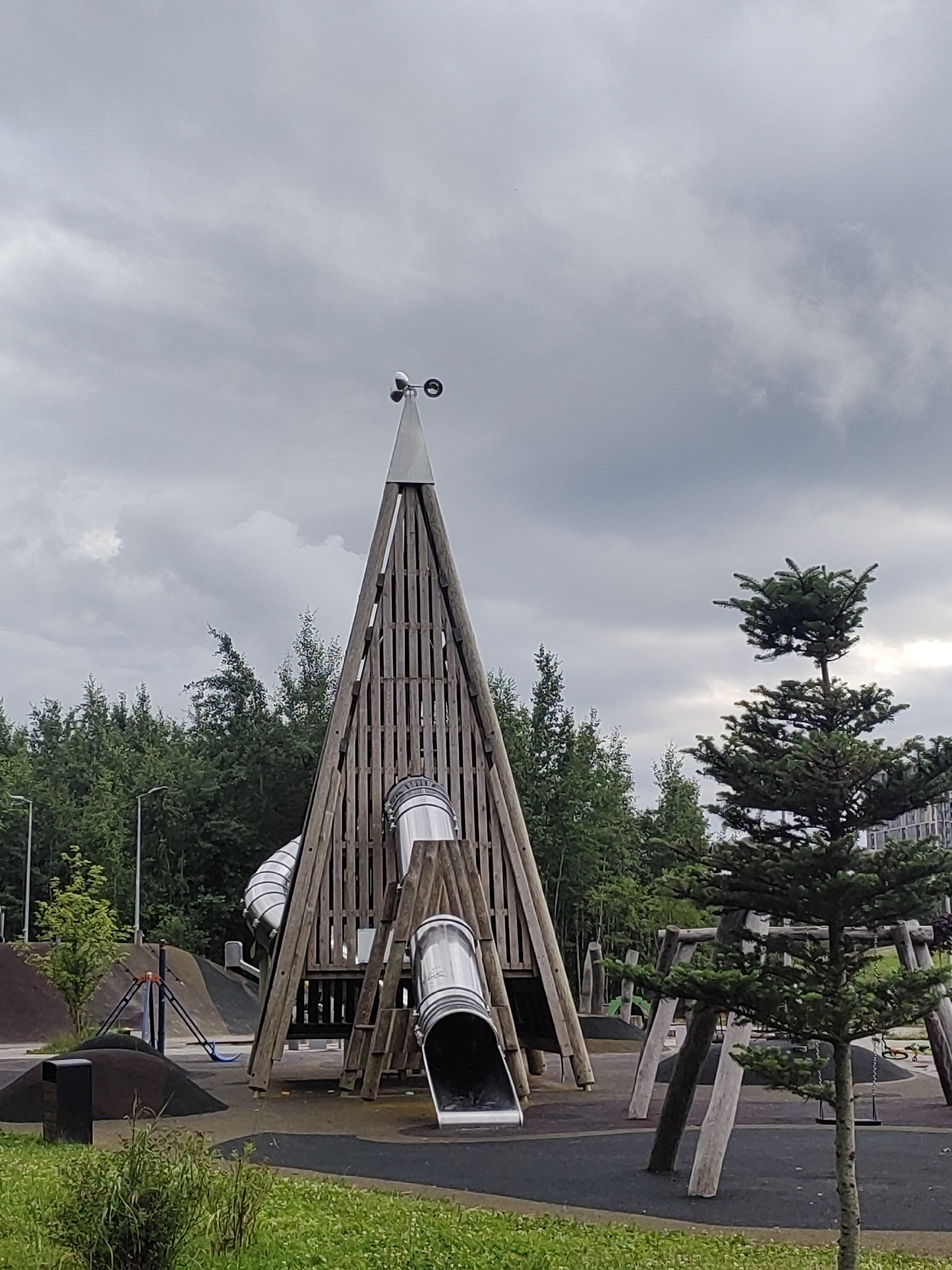 The image depicts an outdoor playground on a cloudy day. The central object is a large wooden play structure with a conical roof, featuring two metal tube slides. A wind vane is mounted on top of the structure. To the right, a small evergreen tree stands in front of a more distant, larger evergreen tree, and behind them, a building with multiple floors is partially visible. In the background, a dense line of deciduous trees forms the edge of the playground. The ground is a mix of dark rubber surfacing and green grass. The scene appears to be located in a park or residential area within the city of Saint Petersburg, Russia, likely in a modern housing development given the presence of the building. There are no people visible in the image.