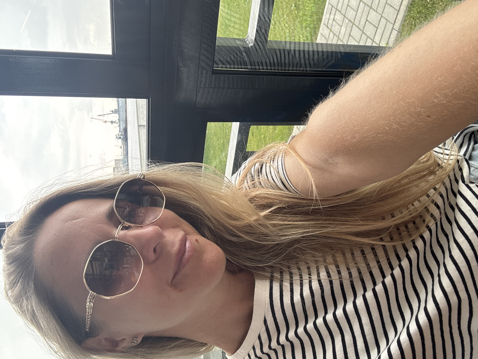 A blonde woman in her 30s with long, wavy hair and sunglasses is taking a selfie. She is wearing a black and white striped t-shirt. She is seated indoors, with a large window behind her that offers a view of a port with cranes and ships. A grassy area with paved walkways is visible outside the window. The lighting suggests it is daytime with an overcast sky. The exact location in Chaikovsky, Russia, cannot be determined from the image.