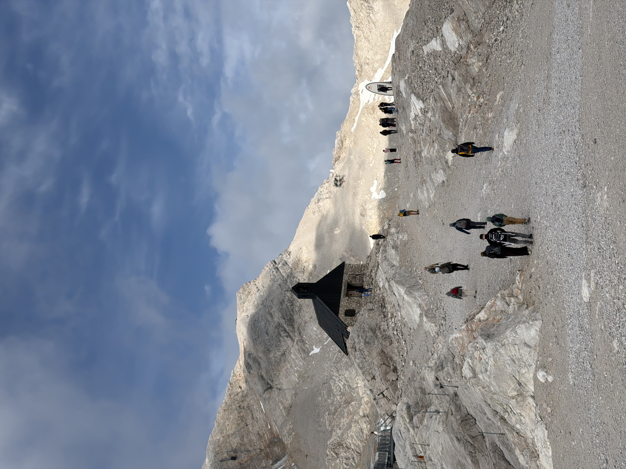The image depicts a mountainous landscape with a small, dark-roofed building resembling a chapel situated on a rocky incline. A group of people are scattered across the scene, walking on the gravelly terrain. Some are walking towards the chapel, while others are moving away. The environment is characterized by rugged, light-colored rocks and patches of snow or ice. The sky above is partly cloudy with a mix of blue sky and white clouds. In the background, more mountains are visible, with some appearing to have a ski lift structure. One of the hints shows a mirrored object standing upright in the distance. The overall scene suggests a tourist destination in a high-altitude, alpine setting. Given the context of Garmisch-Partenkirchen, Germany, this location is likely the Zugspitze, Germany's highest peak, known for its chapel and visitor facilities.