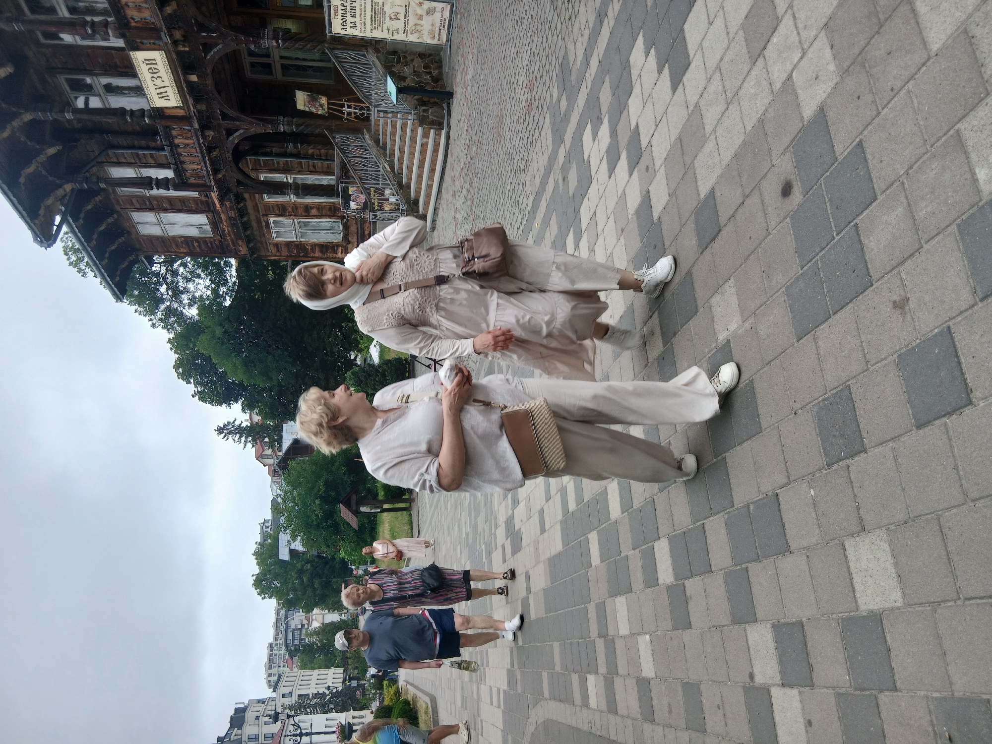 In Truskavets, Ukraine, two women are captured in a vertical photograph, standing on a paved walkway. One woman, in the foreground, wears a light-colored headscarf and a matching loose-fitting outfit with a cross-body bag. The other woman, positioned slightly behind and to her left, is also dressed in a similar light-colored, loose-fitting ensemble.
Behind them, a historic wooden building with intricate carvings and a sign reading "МУЗЕЙ" (Museum) in Cyrillic is visible, suggesting a cultural or historical site. The walkway is constructed with interlocking gray and beige pavers. In the background, trees and a cluster of buildings indicate a town or city setting. The sky appears overcast, suggesting a cloudy day. The overall mood seems relaxed and casual, possibly a sightseeing outing. Other individuals are partially visible in the background, also walking on the paved area.