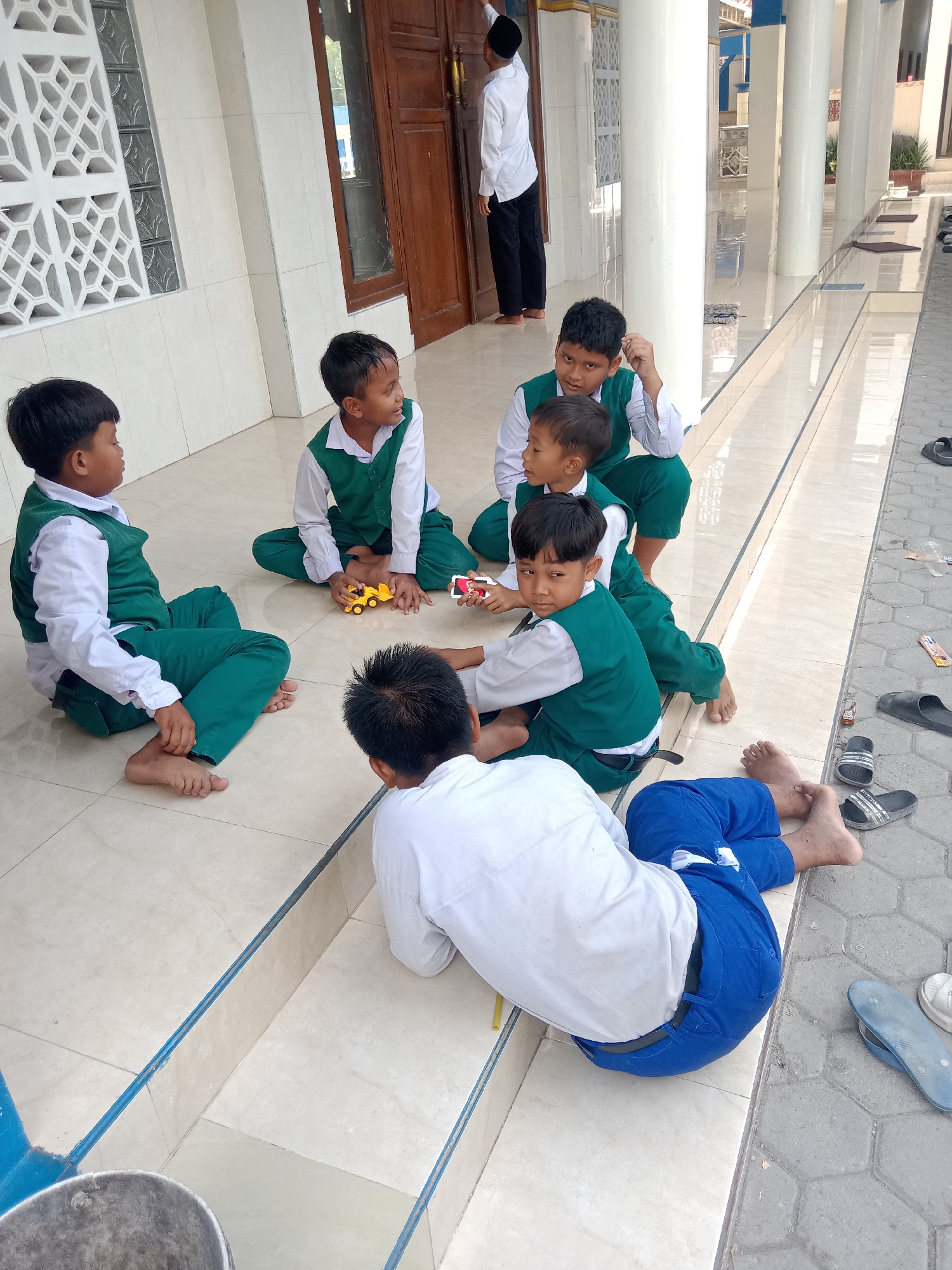 In Sugihwaras, Indonesia, a group of young boys, likely students, are gathered on the tiled steps and ground outside a building with a large wooden door. The boys are wearing green vests over white shirts and green pants, suggesting a school uniform. Some are sitting on the ground, while others are on the steps. One boy in the foreground is lying on his side, wearing blue pants and a white shirt. Another boy is holding a small yellow toy car. In the background, a man in a white shirt and black pants, wearing a black cap, is standing near the door. The environment appears to be an outdoor area with a tiled walkway, pillars, and decorative window features on the building. Several pairs of sandals are scattered on the ground near the walkway.