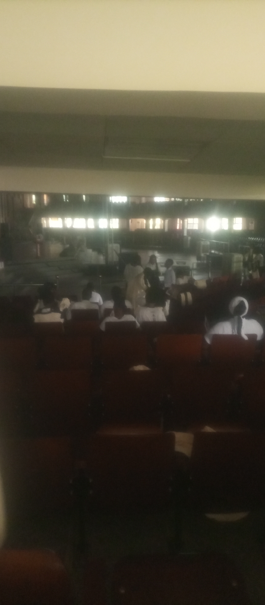 The image depicts an indoor gathering, possibly in a school or community hall in Kuson, Nigeria. In the foreground, rows of dark brown chairs are occupied by individuals, many wearing white attire. The lighting is dim, with bright windows visible in the background across a large, open space. Several people are standing and interacting in the mid-ground, some appearing to be listening to someone or observing an event. The overall impression is one of a formal or religious assembly.