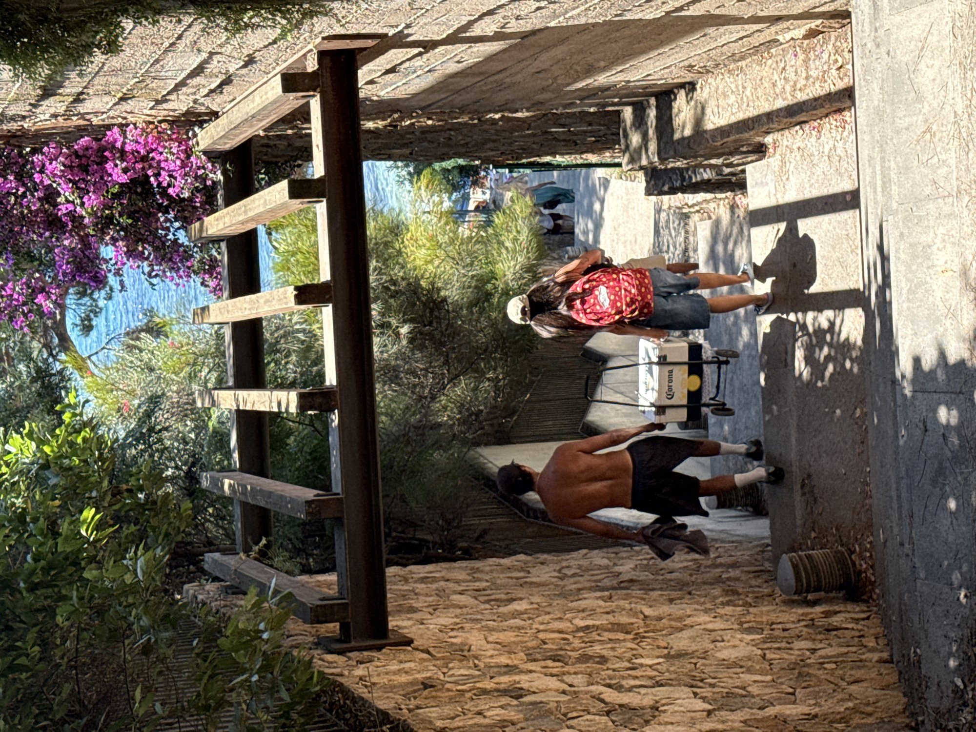 Two individuals descend a stone staircase surrounded by vegetation and a wooden overhead structure. On the left, a shirtless man wearing dark shorts and white socks carries a folded dark cloth in his left hand. To his right, a person with a baseball cap and a red backpack with white polka dots walks down the stairs, wearing denim shorts and white shoes. They are walking behind a white cooler with a yellow "Corona" logo and wheels. The staircase leads towards a bright, sunlit area with the blue sea visible in the background, framed by flowering bushes. The stone walls of the staircase are textured and appear aged. The scene suggests a path leading to or from a coastal area in Castell d'Aro, Platja d'Aro i s'Agaró, Spain, likely on a sunny day.